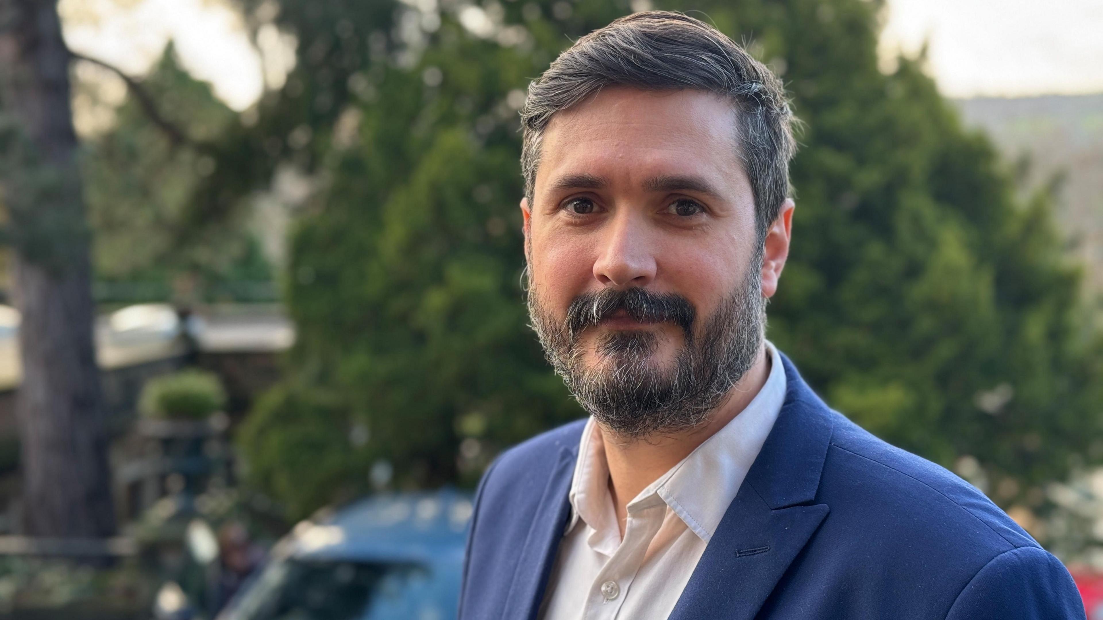 Stephen Reed of Derbyshire County Council (beard, full head of dark hair with streaks of grey) standing outdoors wearing a blue jacket with a white open neck shirt.