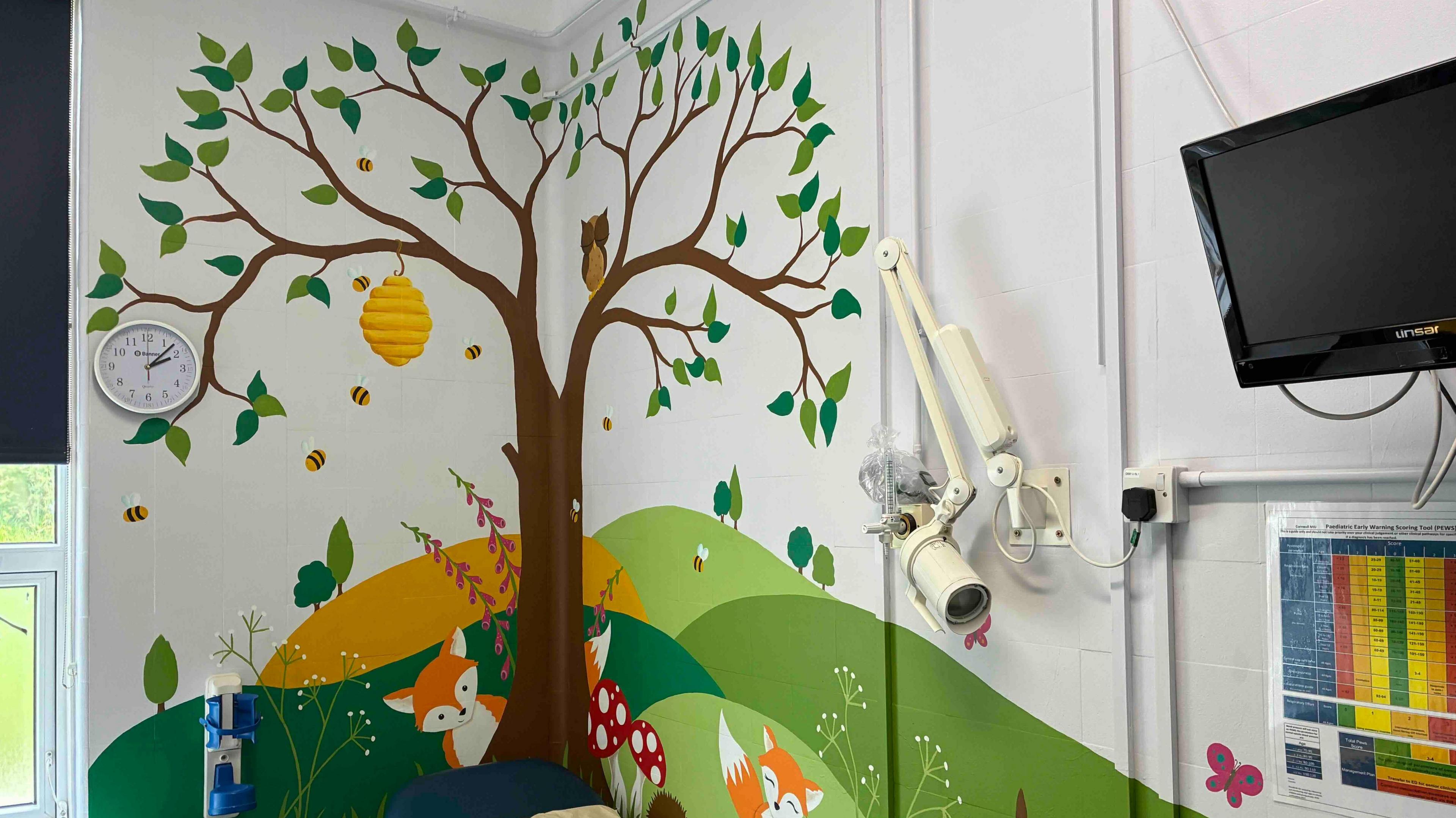 A mural of a tree with animals and wildlife with some equipment and a television on the wall.