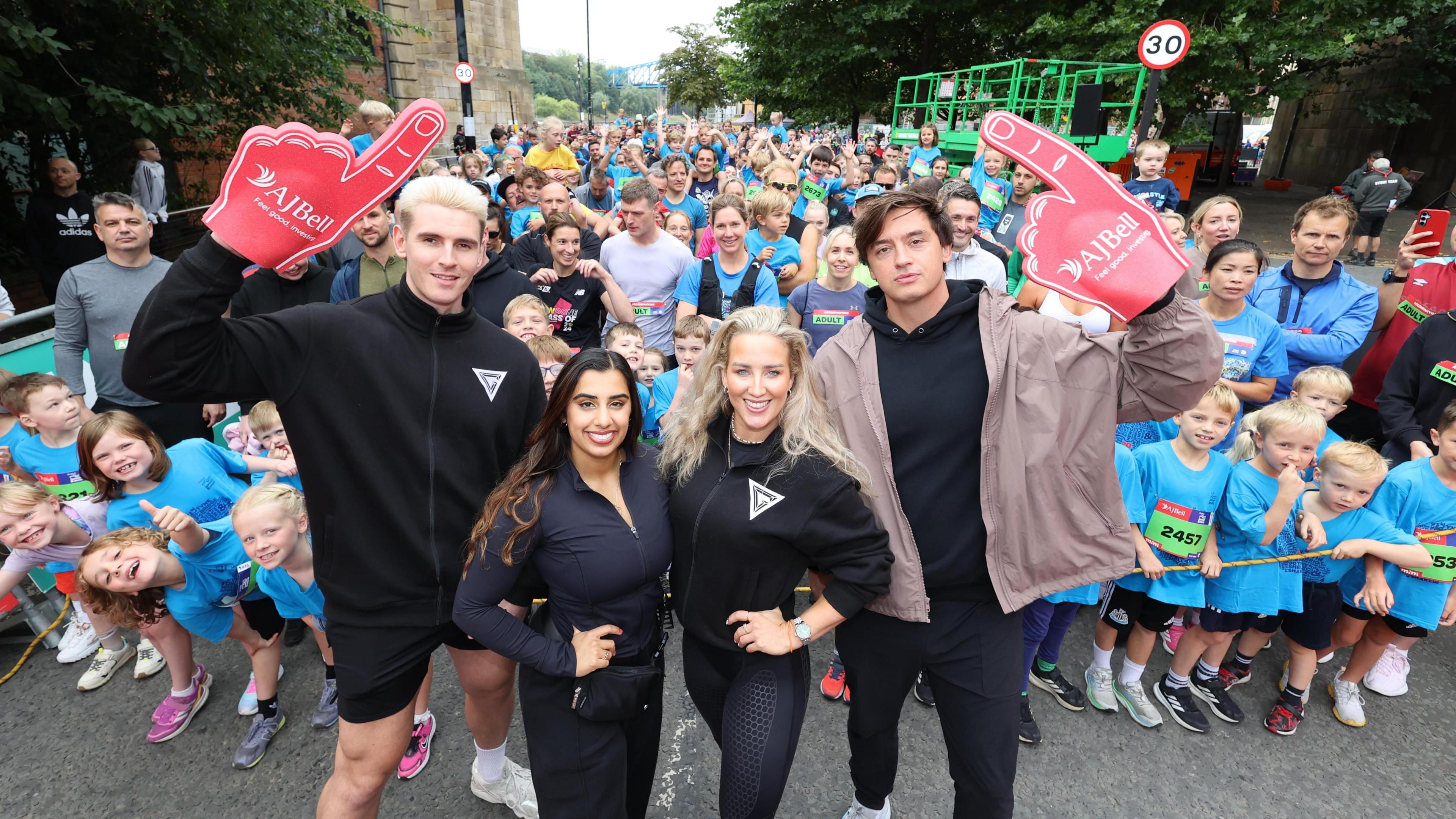 Bionic, Athena, Sabre and Apollo, who are wearing all black, stand in front of a group of children posing for the camera and standing behind a rope. They are all wearing blue T-shirts and green signs on their fronts with their race numbers.