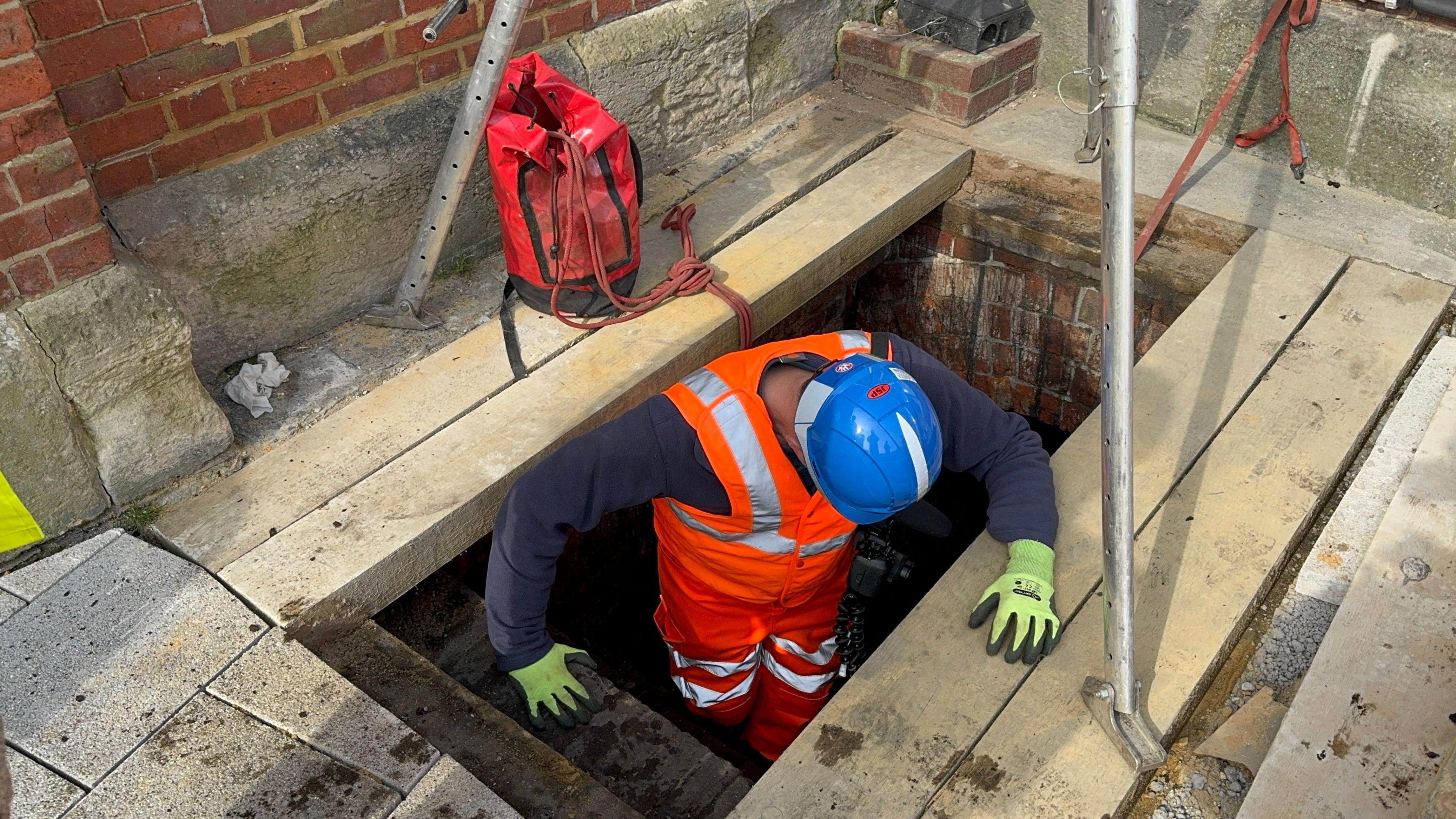 A man wearing an orange hi-vis suit, yellow gloves and a blue hard helmet. He is carefully stepping down a ladder and descending into a dark hole in the ground, which is covered by wooden sleeper blocks. 