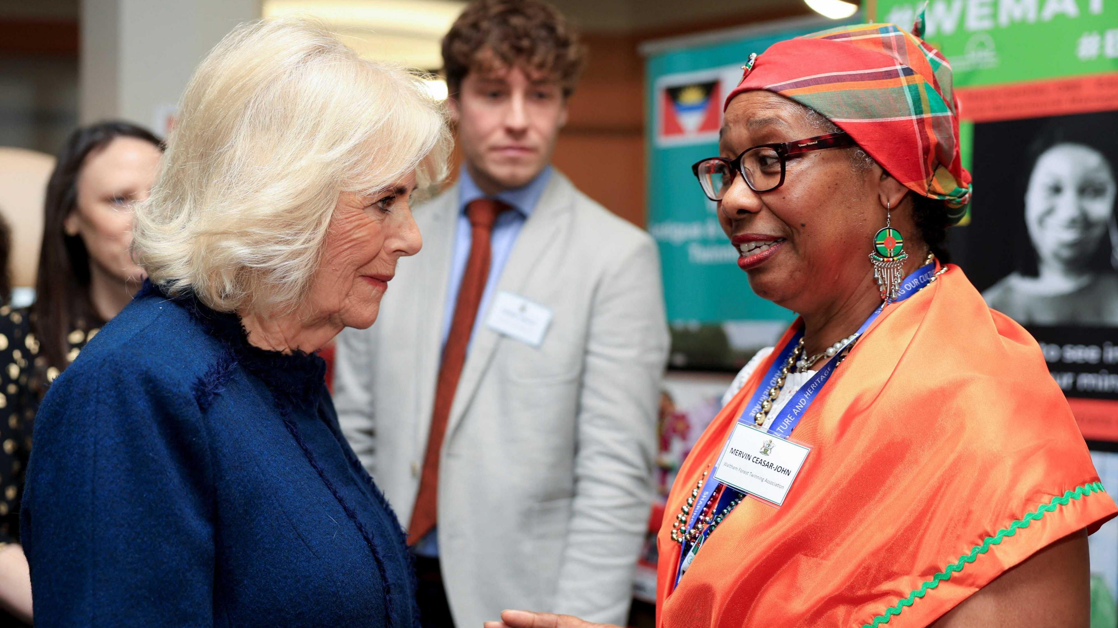 The Queen, in a dark blue dress, speaks to a woman who is wearing a bright orange dress and a multi-coloured headscart. In the background people look on.  