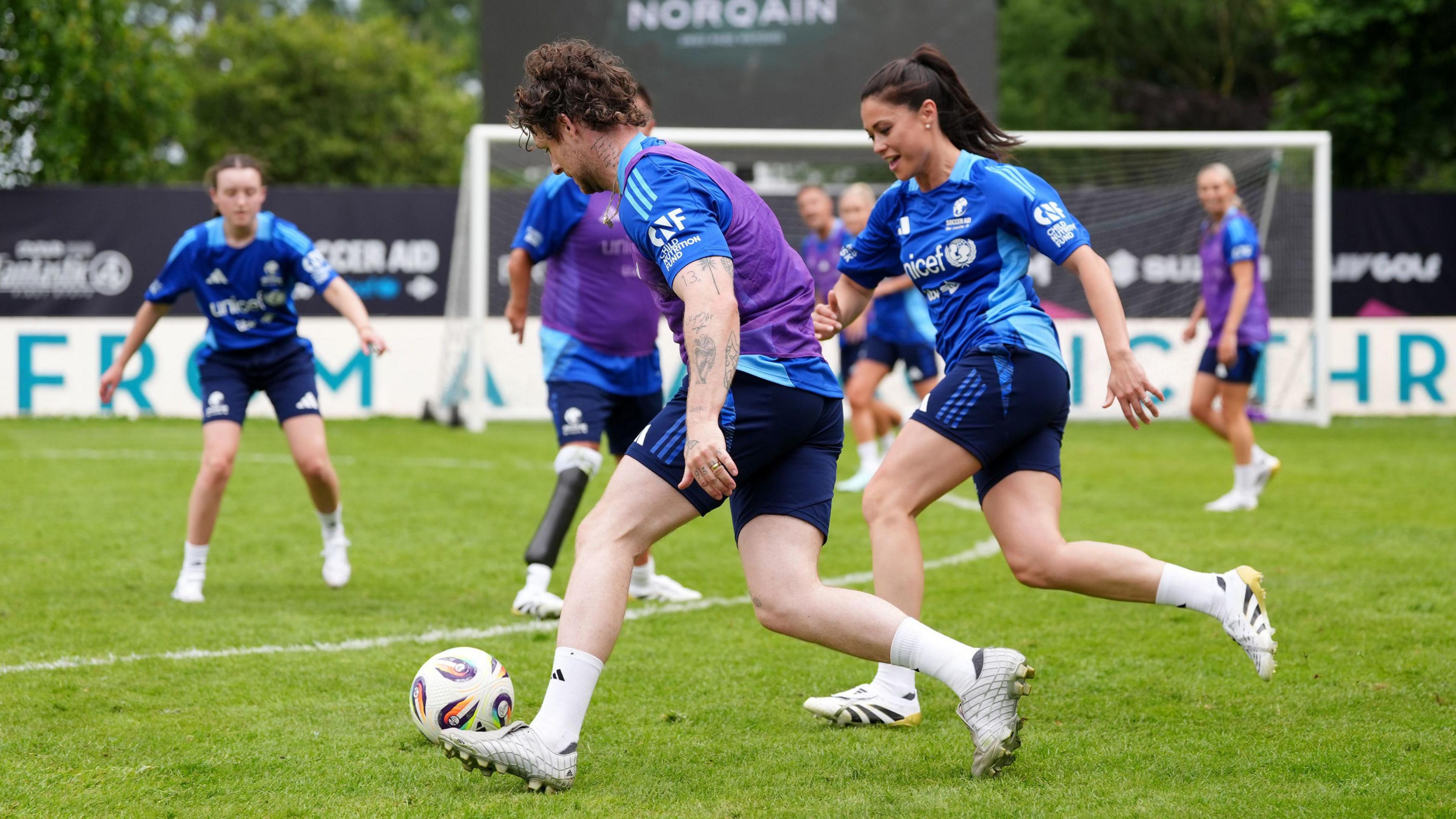 A man dressed in a blue, purple and black football kit, dribbles with a ball past a woman wearing a similar kit. Other players are watching on in the background with a goal visible behind them.