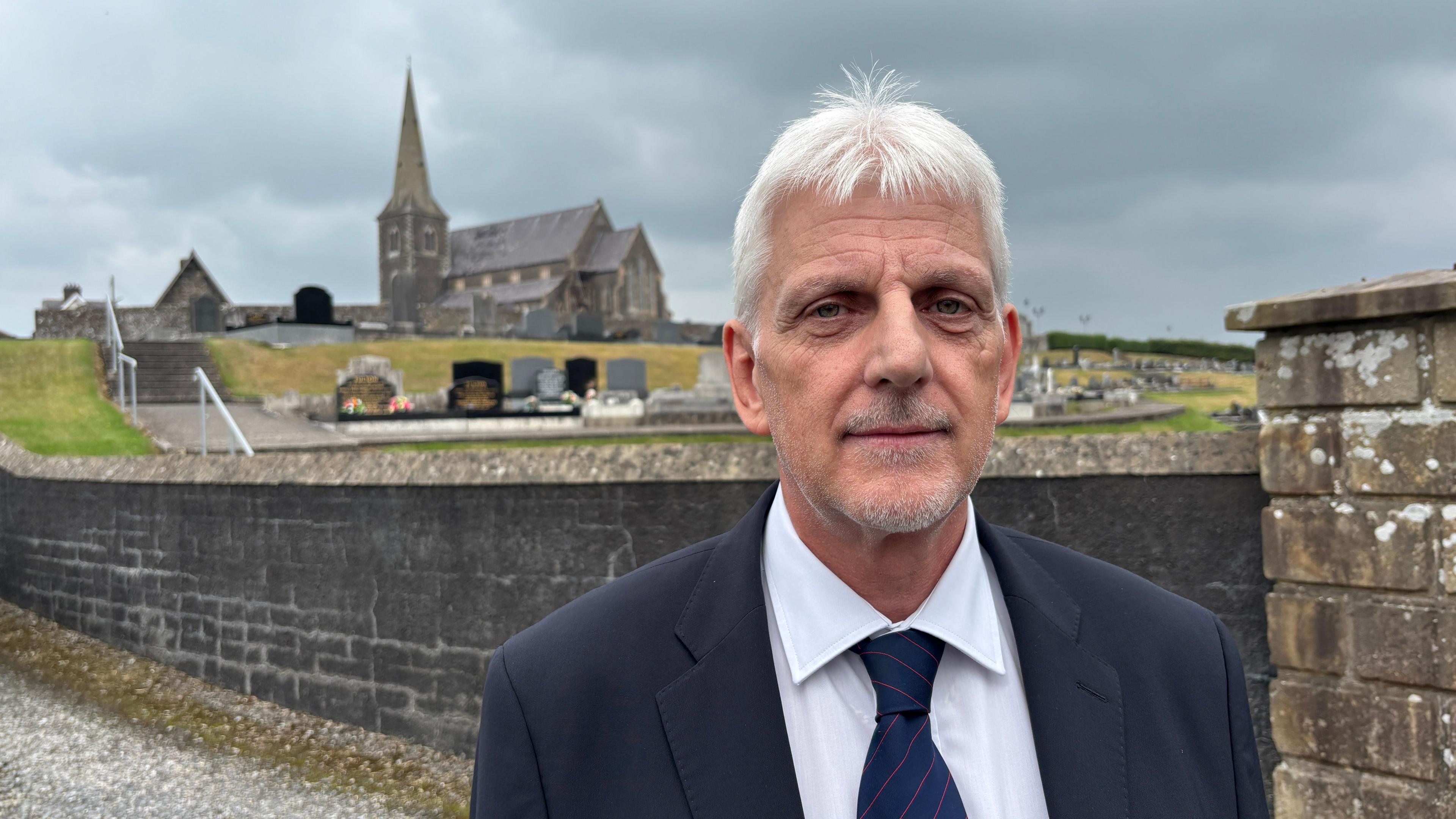 Nigel Dawson is wearing a navy suit with a white shirt and a navy and red striped tie. He's outside and a church is behind him. The sky is cloudy.