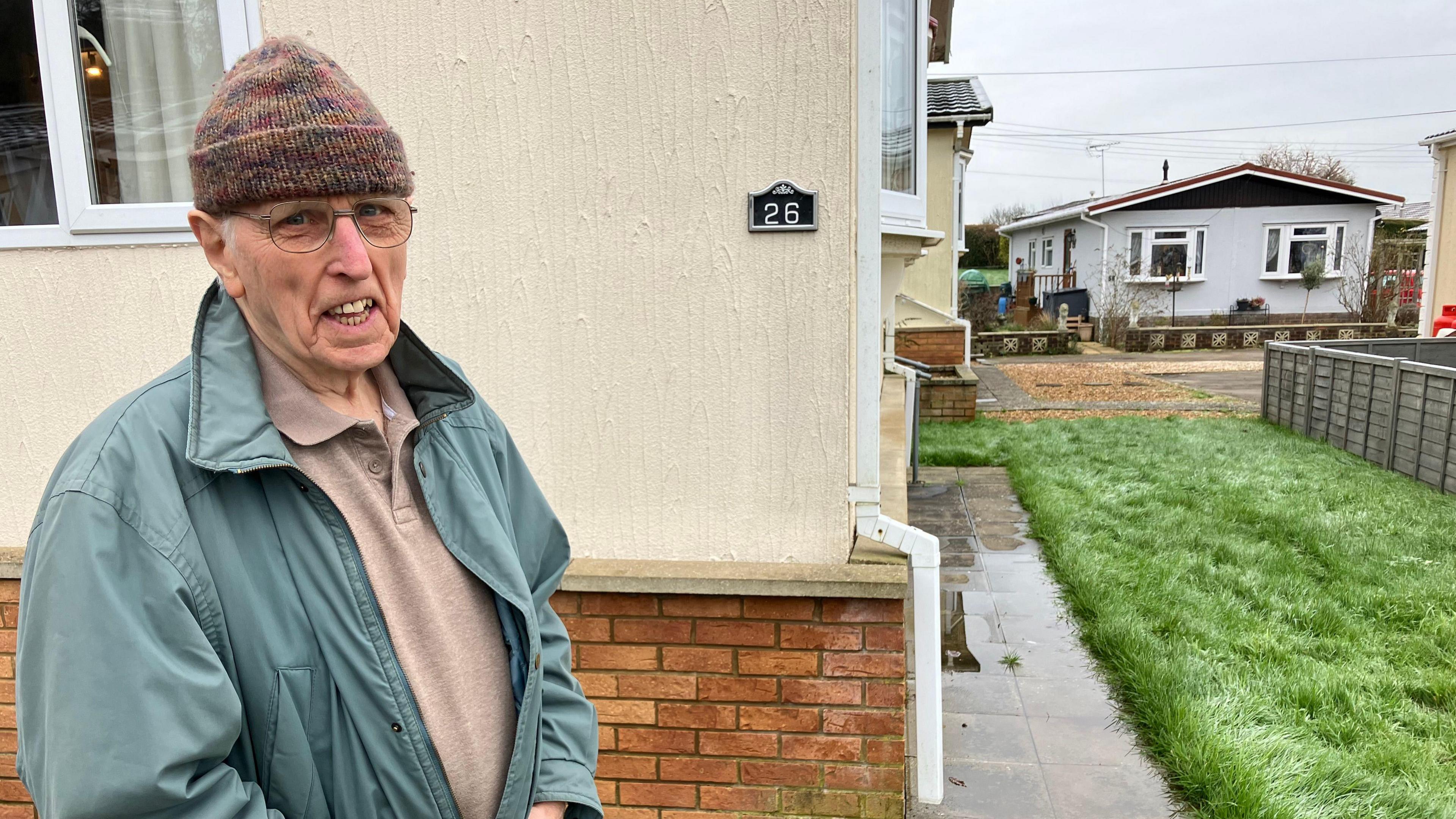 David McGovern, wearing a brown polo shirt, a blue coat stands and a brown wollen hat, stands next to his property. A path on the right hand side is covered in water.