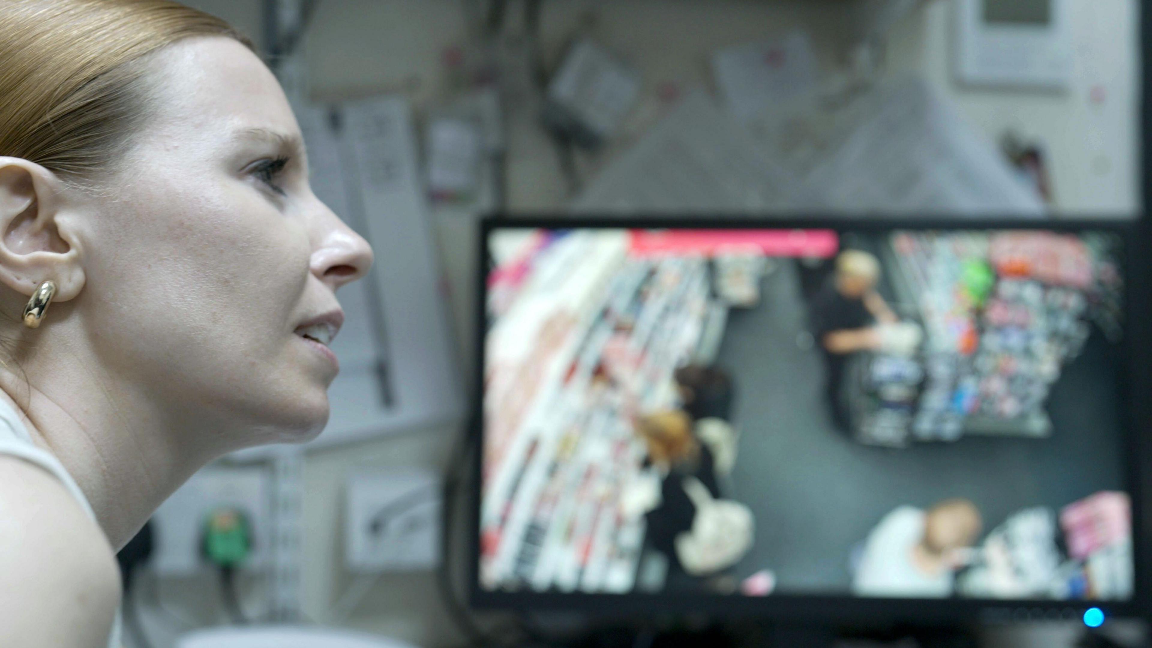 Stacey Dooley sits in front of a CCTV camera. A woman with ginger hair and a gold hoop earring sits in front of a CCTV camera, which shows footage of a shop floor. 