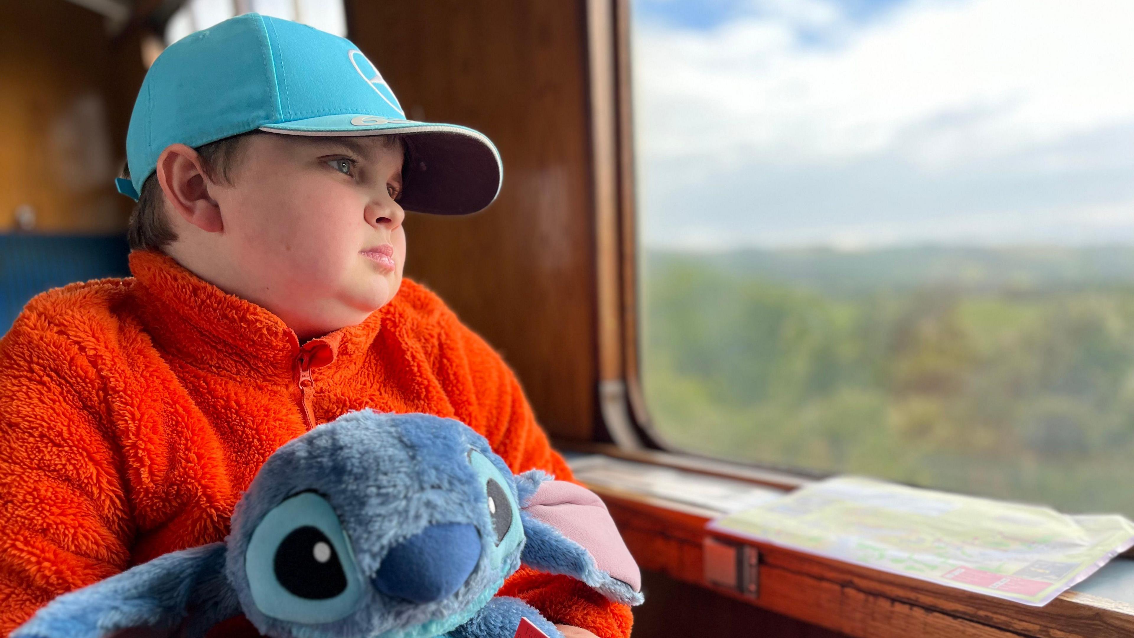 Boy wearing blue hat, orange fleece and holding blue teddy looking out of train window.