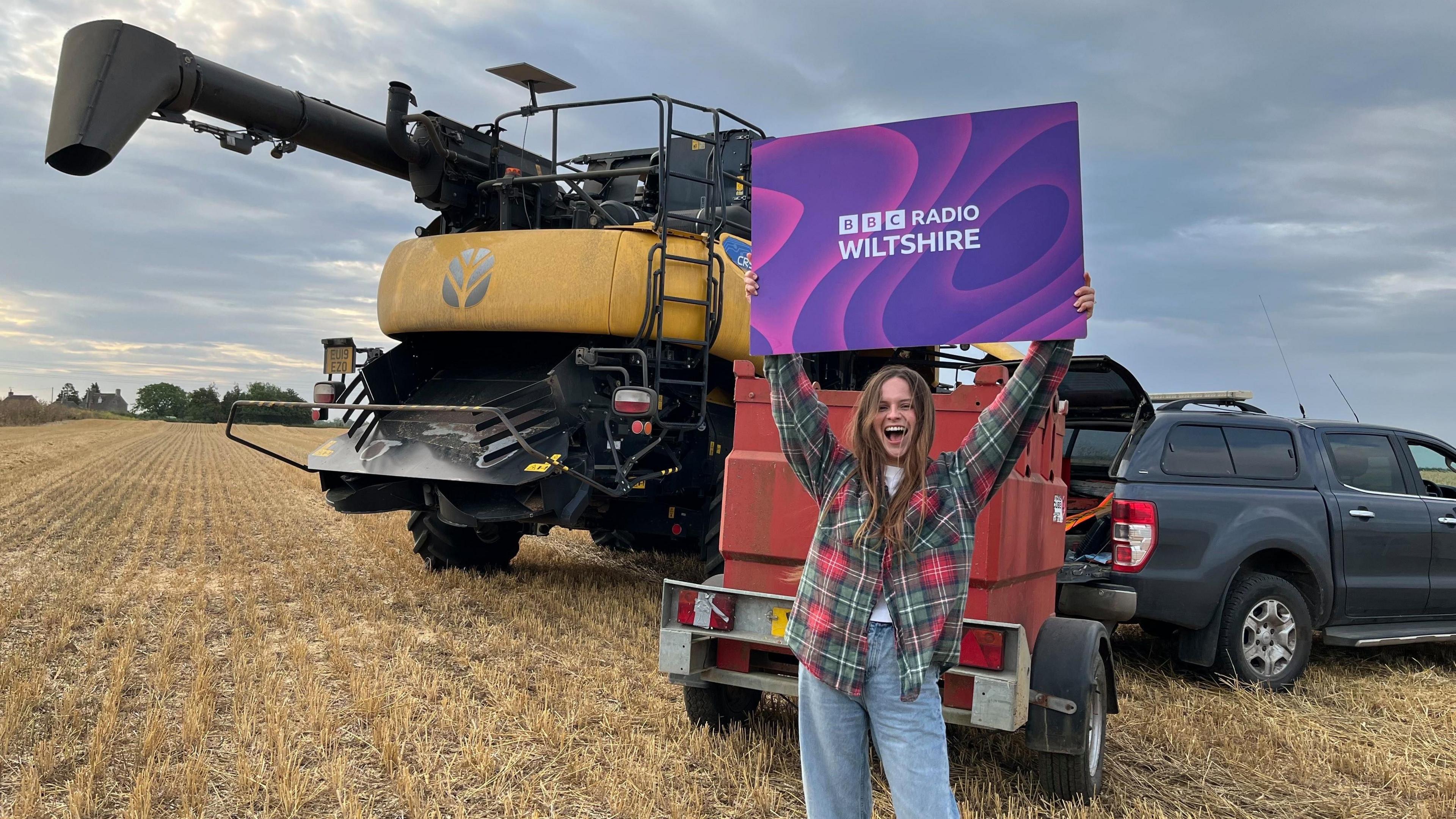 Farmwatch Gabrielle Aplin singer and sheep farmer holds a purple sign saying 'BBC Radio Wiltshire' in front of farming equipment