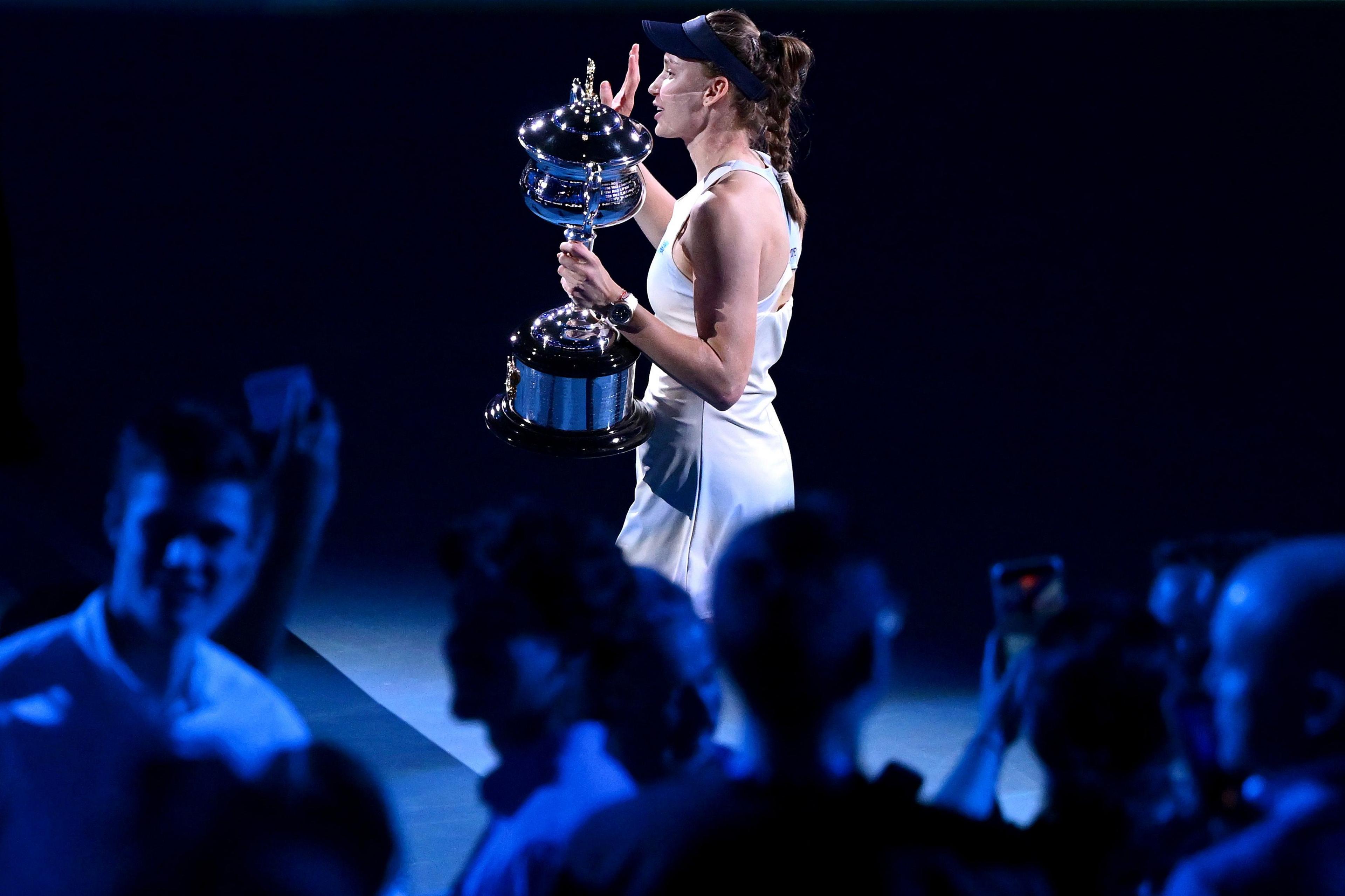 Tennis player Elena Rybakina, wearing a white dress, holds a large silver trophy aloft and acknowledges the fans after winning the Australian Open with victory over Aryna Sabalenka.
