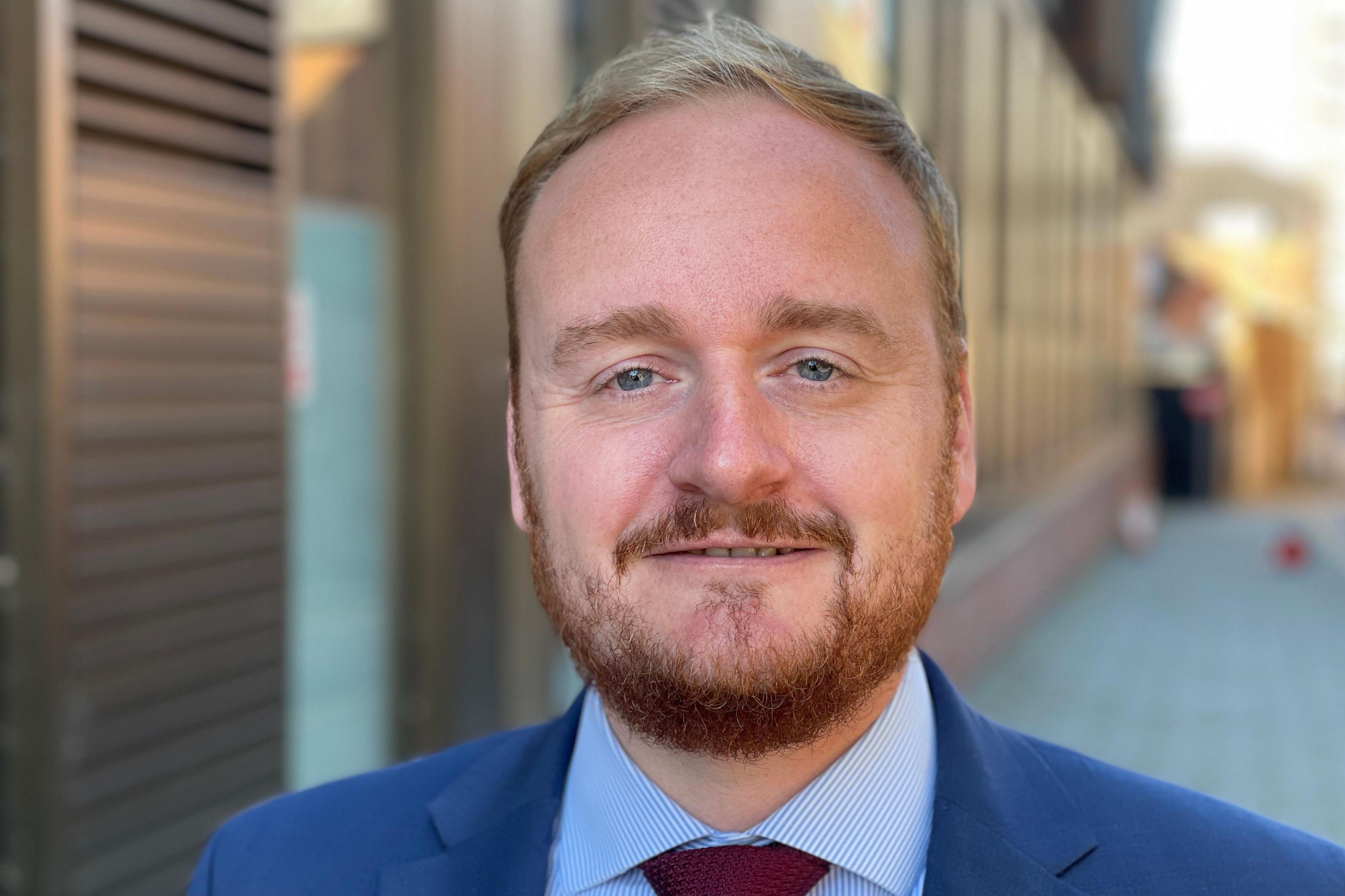 Adam Brown is wearing a light blue suit with a blue and white pinstriped shirt and dark red tie.