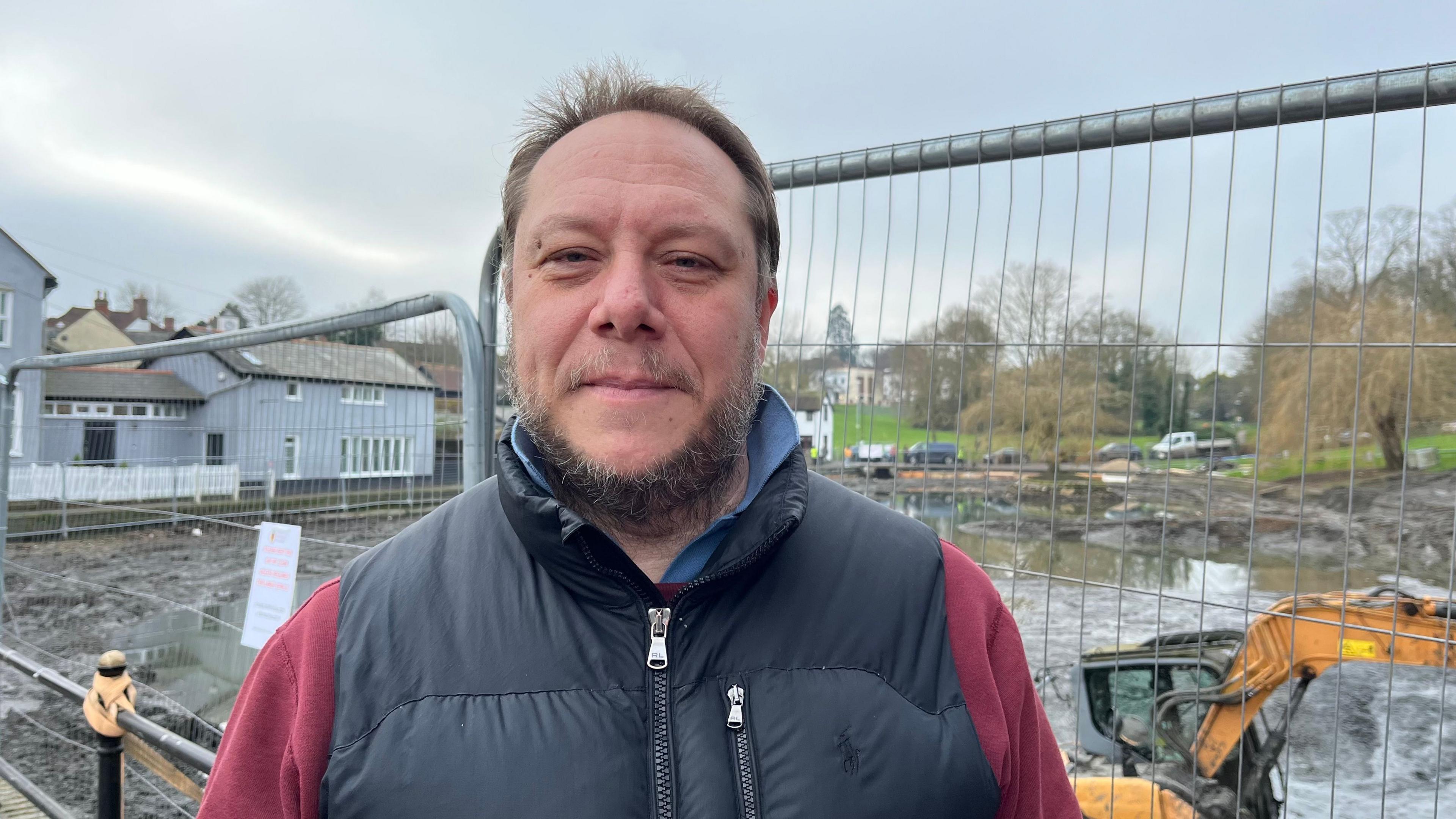 Jonathan Little wearing a black gilet and maroon jumper. He has short brown hair and a beard and is smiling while stood in front of the digger, which is stuck in mud.