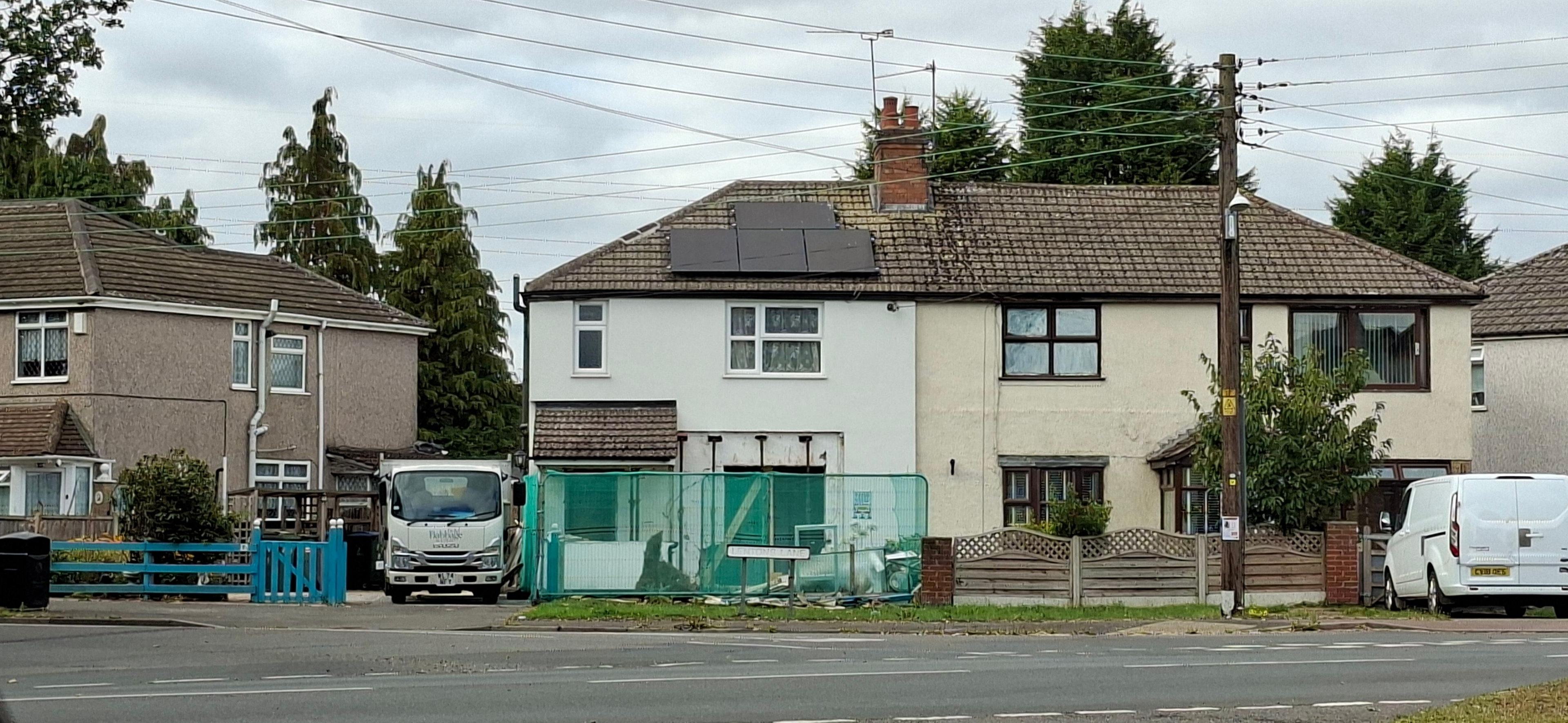 A semi-detached house on a residential street. It is behind a road junction and has a green fence around the front of the house after a car had crashed through the front window into the property. 