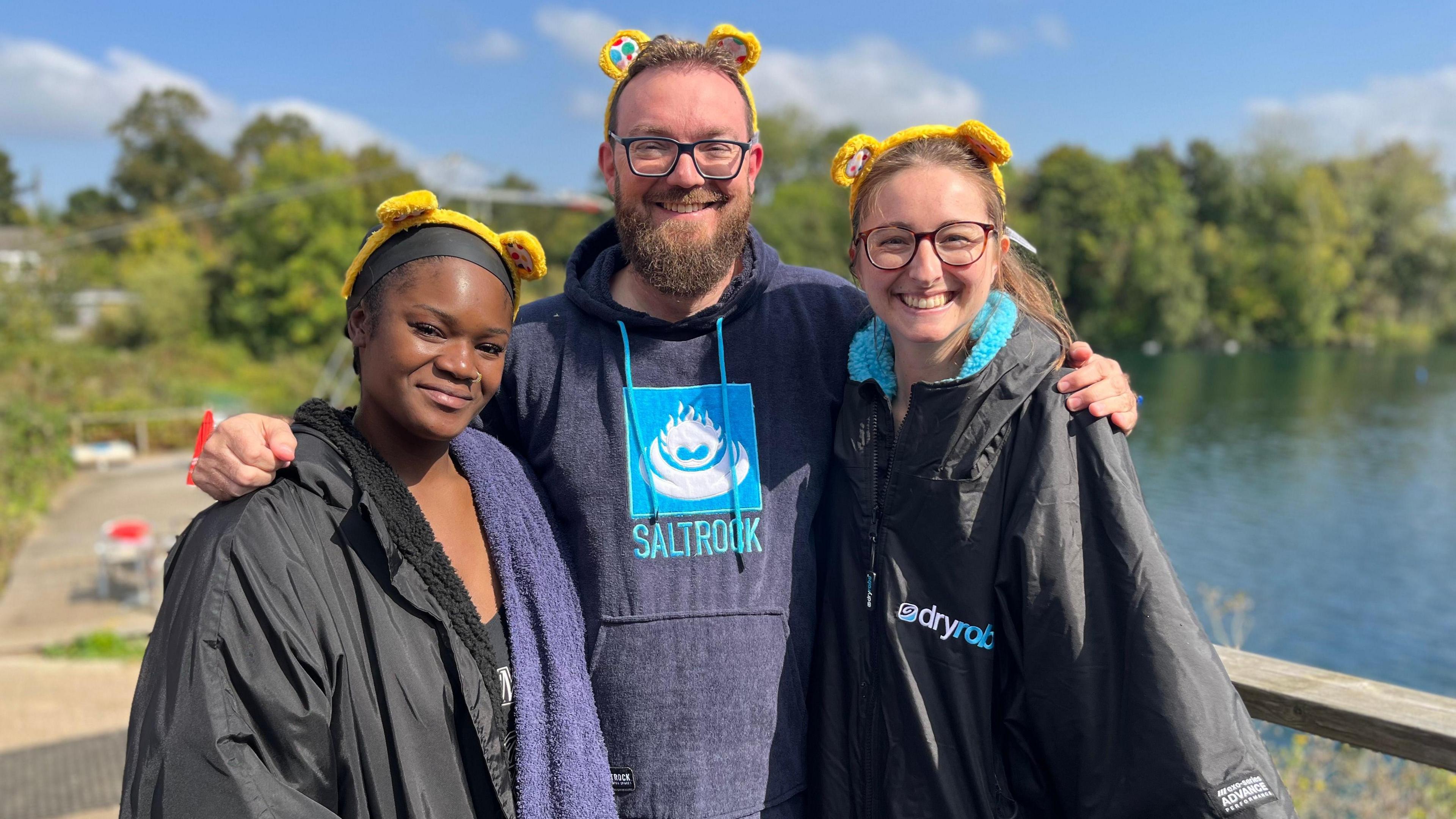 Two women wearing coats and a man wearing a changing towel standing in front of a lake on a sunny day. They're smiling and each wearing a set of yellow Pudsey Bear ears.