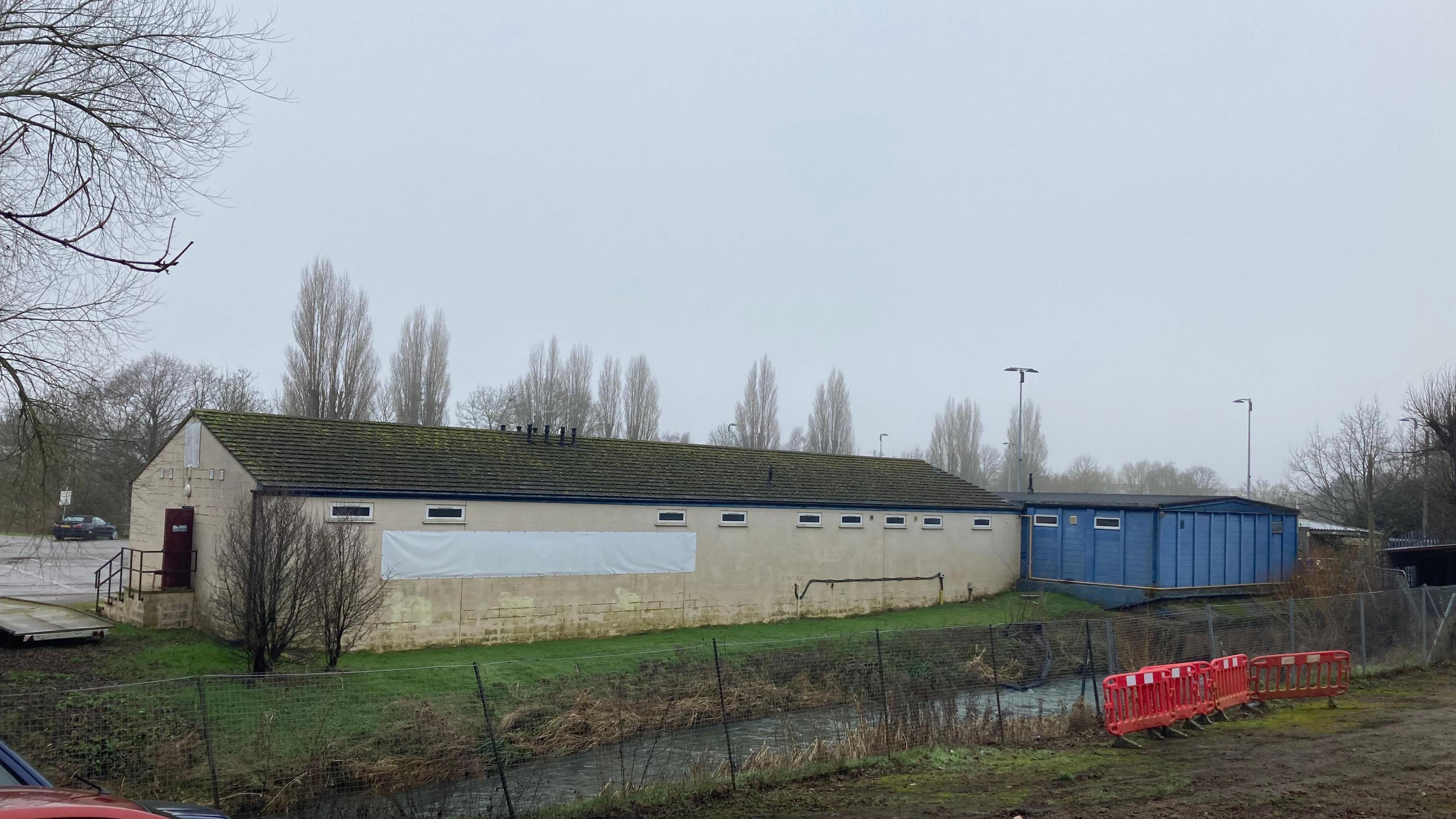 A long brick building with a blue brick building attached at the back. A canal runs along in front of it.