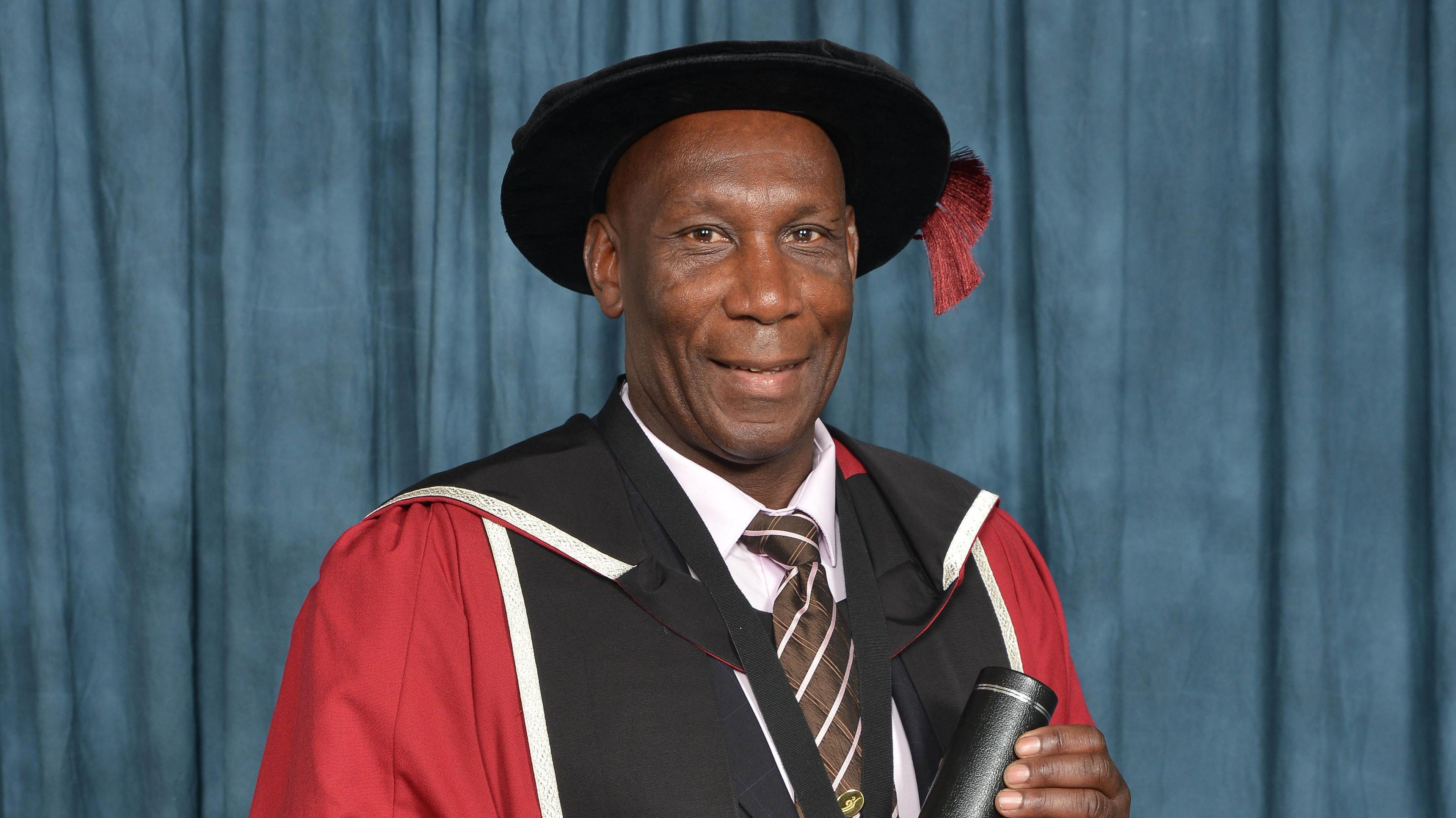 A man is wearing a university cap and gowns and is holding a tube with a university certificate inside