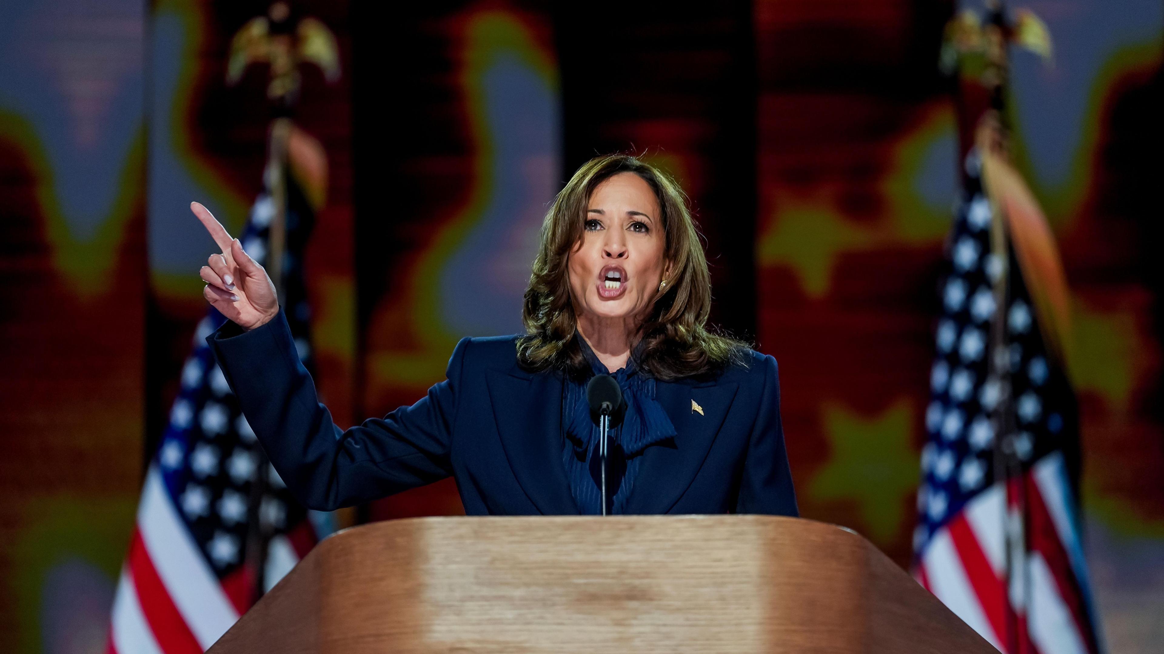 A woman in a navy blue suit points into the air at a wooden podium with two American flags in the background.