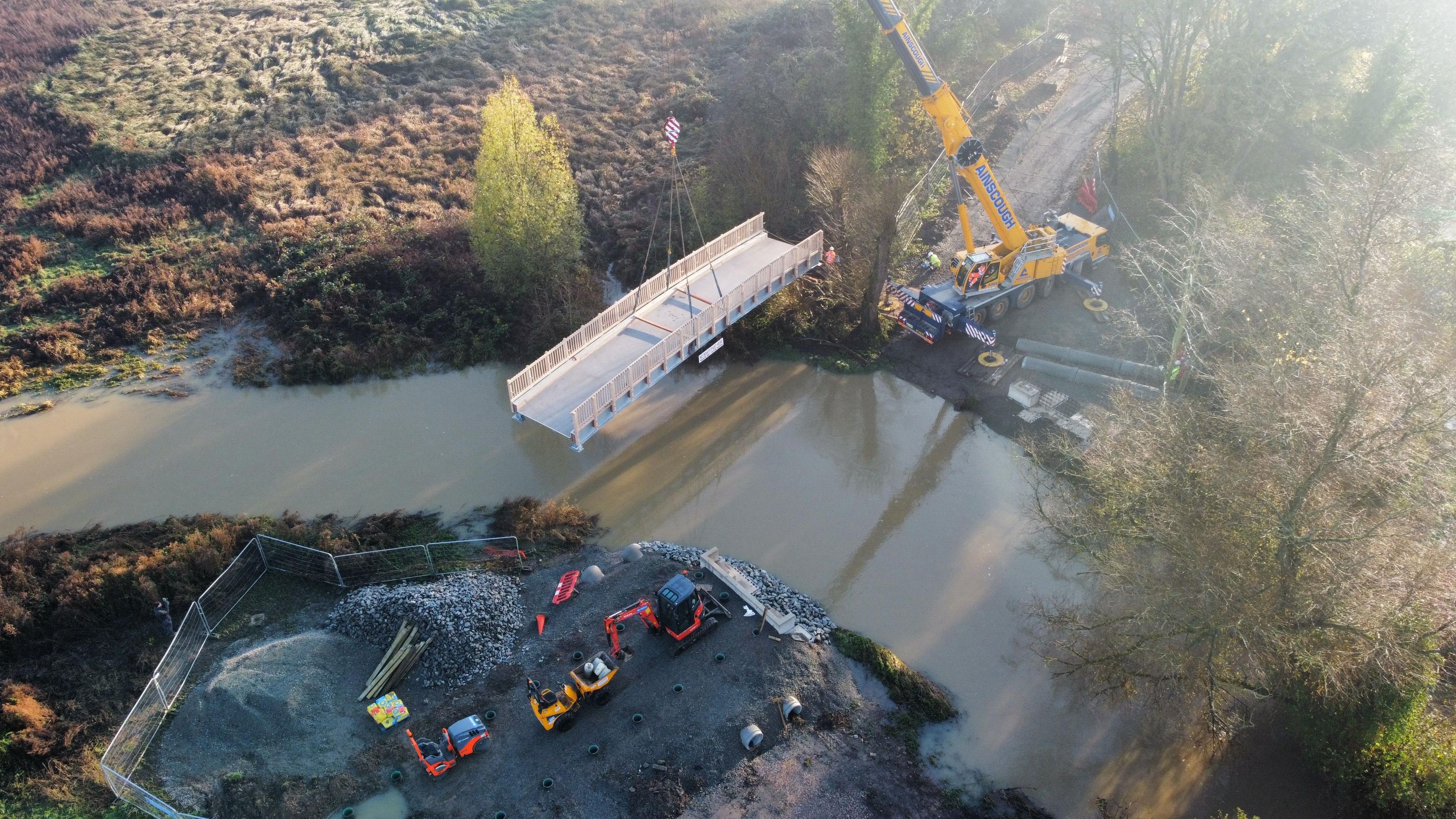 An aerial view of a footbridge over a river being hoisted into place by a yellow crane. People in high-vis yellow and orange jackets and hard hats stand on the far riverbank near the crane. On the opposing closer bank a small red digger can be seen and yellow and red vehicle used to transport waste. There are metal barriers around this area. Trees line the riverbank on the far side, and a grassy field lies to the left. The river has partially burst its banks and water has flooded onto the field.