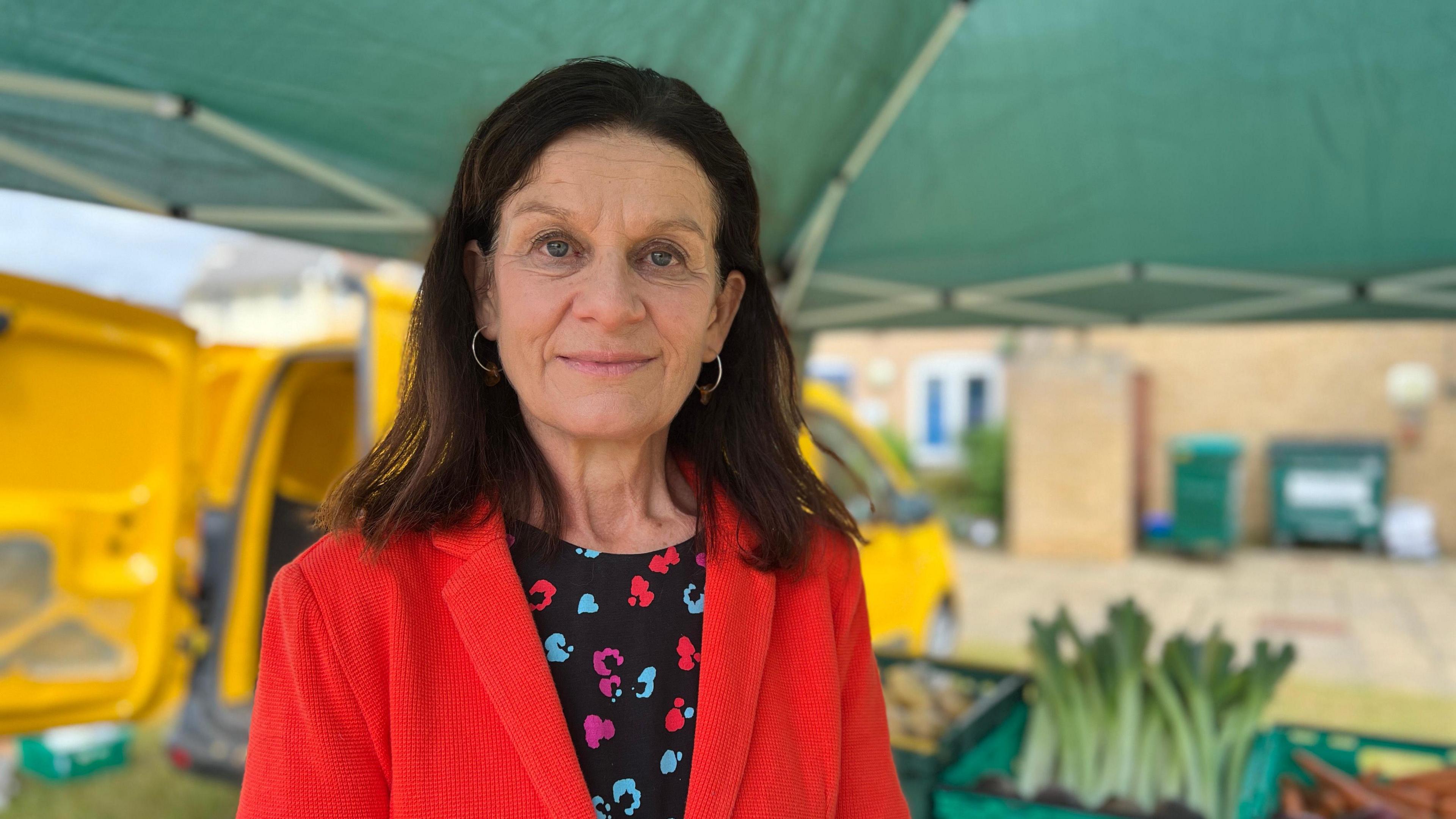 Bridget Smith faces the camera wearing a red jacket and a black top with a purple, blue and red pattern. She stands under a green marquee with vegetables baskets in the background.