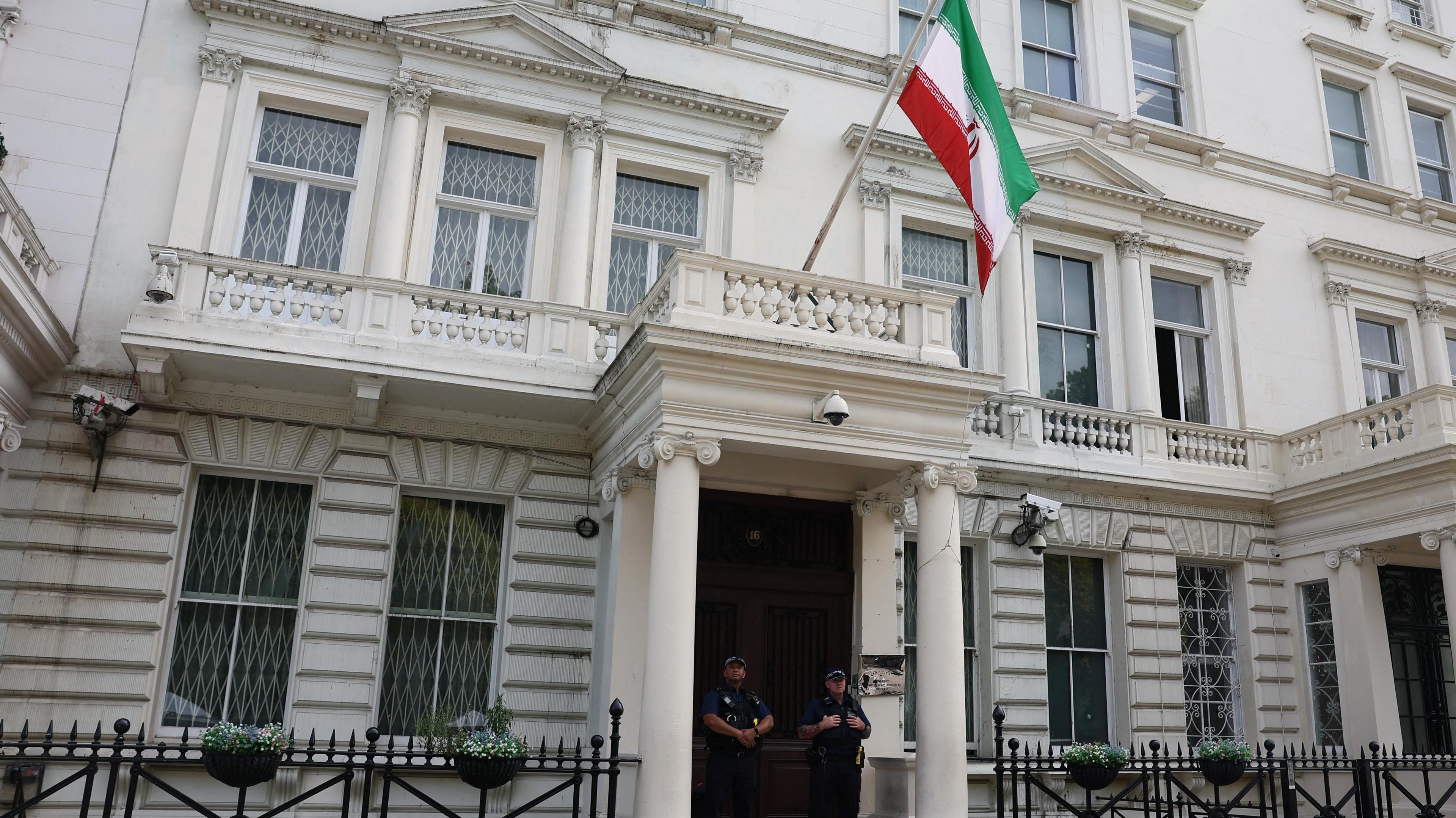 Two police officers stand outside a white Iranian Embassy building flying the Iranian flag, with barred windows and flower baskets on the fence.