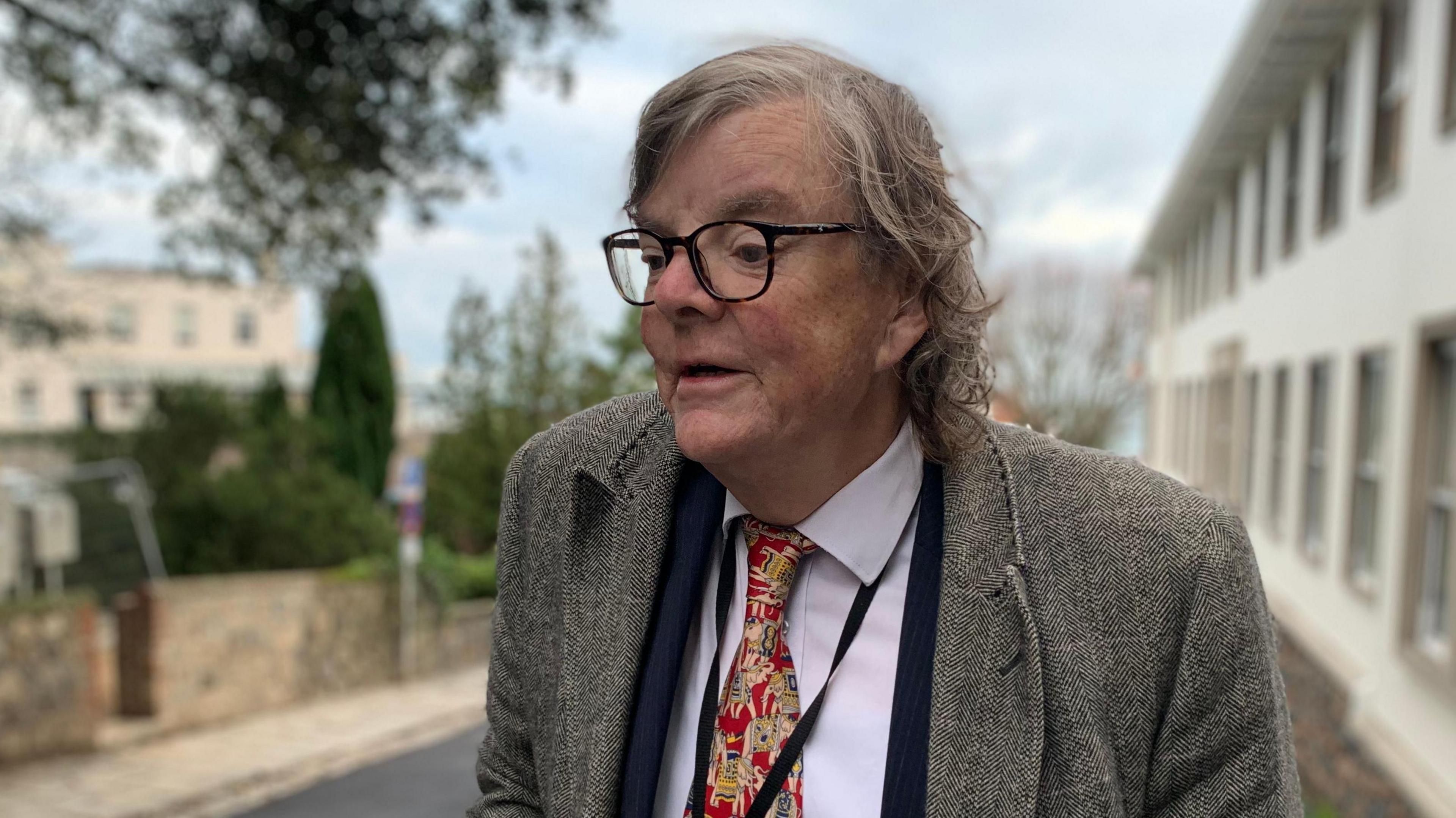 Charles Parkinson standing in the street. He is wearing a grey jacket, a blue suit jacket, a white shirt and a patterned red, white and yellow tie. He has medium length curly hair and thick rimmed black glasses. The street in the background is blurred. 