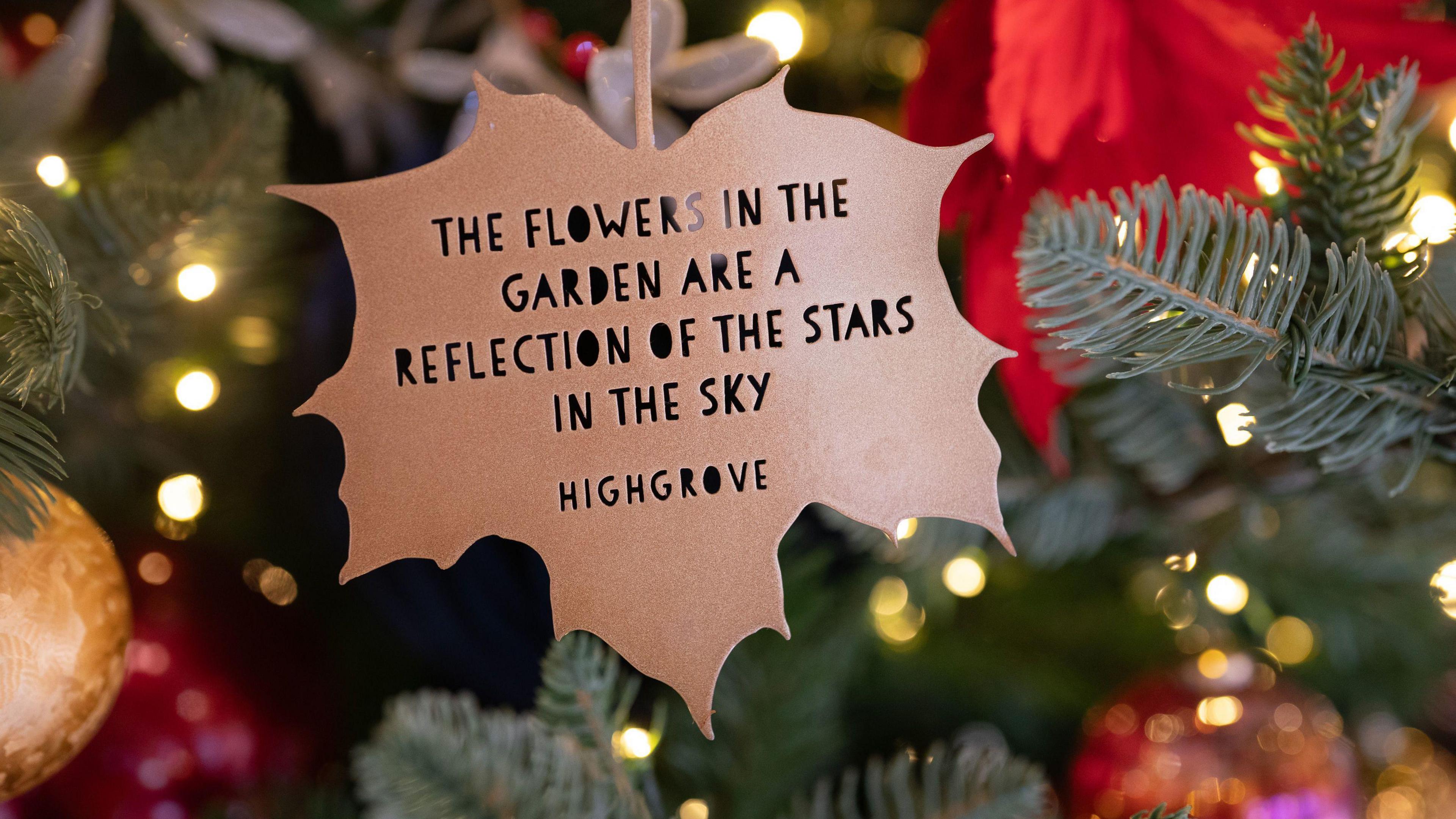 A bronze leaf shaped Christmas tree decoration hangs from a branch, surrounded by fairy lights and gold and red decorations. The leaf reads: "The flowers in the garden are a reflection of the stars in the sky. Highgrove."