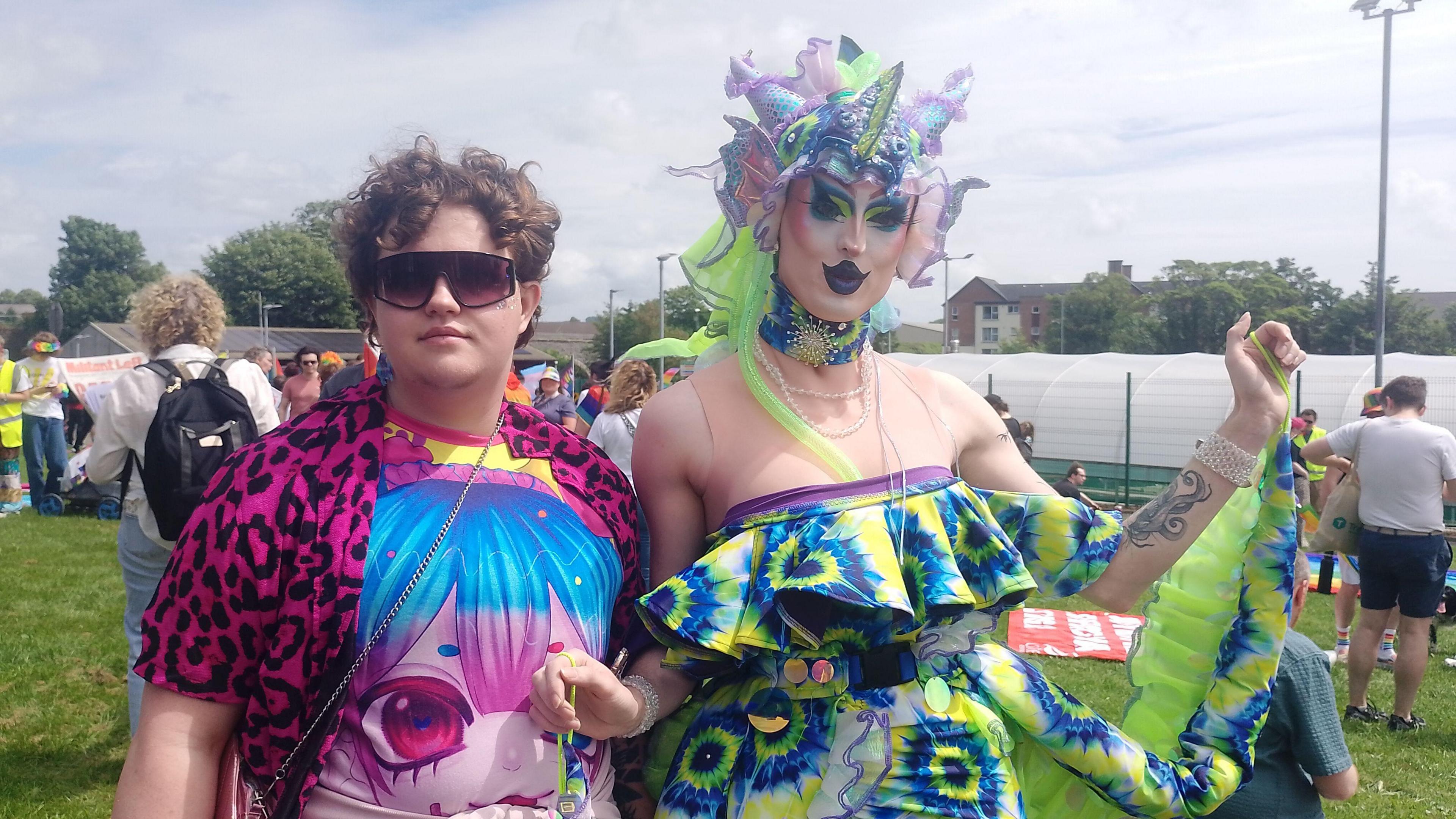 Espresso Martini and Coral Hole Mandi. Espresso Martini has short brown curly hair, wearing sunglasses, a leopard print pink shirt and bright coloured t-shirt. Coral Hall Mandy is wearing a fish-themed headpiece, bright makeup and multi-coloured dress.