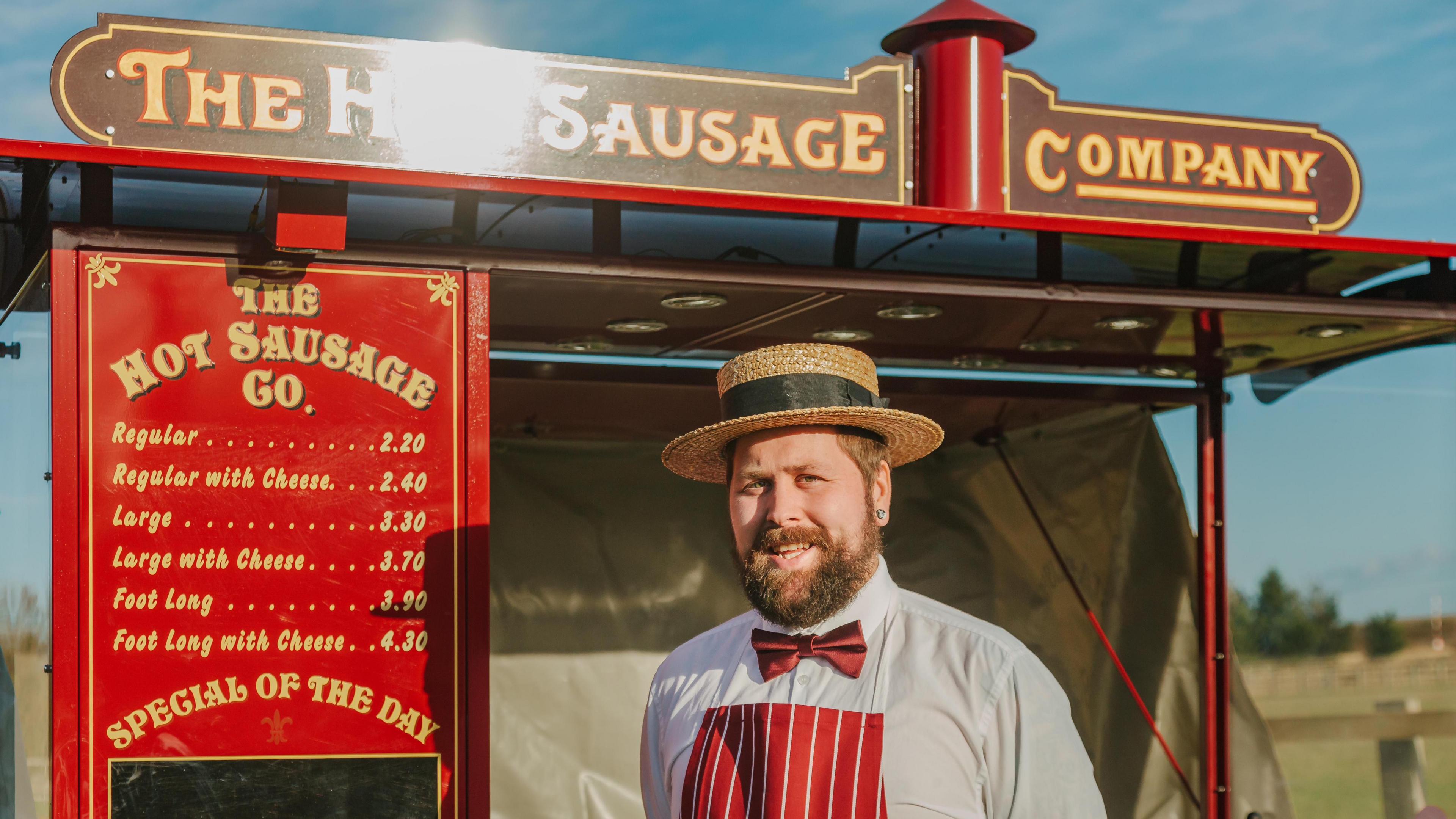 Joshua Bavington-Barber stands in front of his hot dog stand. He wears a straw bowler hat, white shirt, red bow tie and red apron.