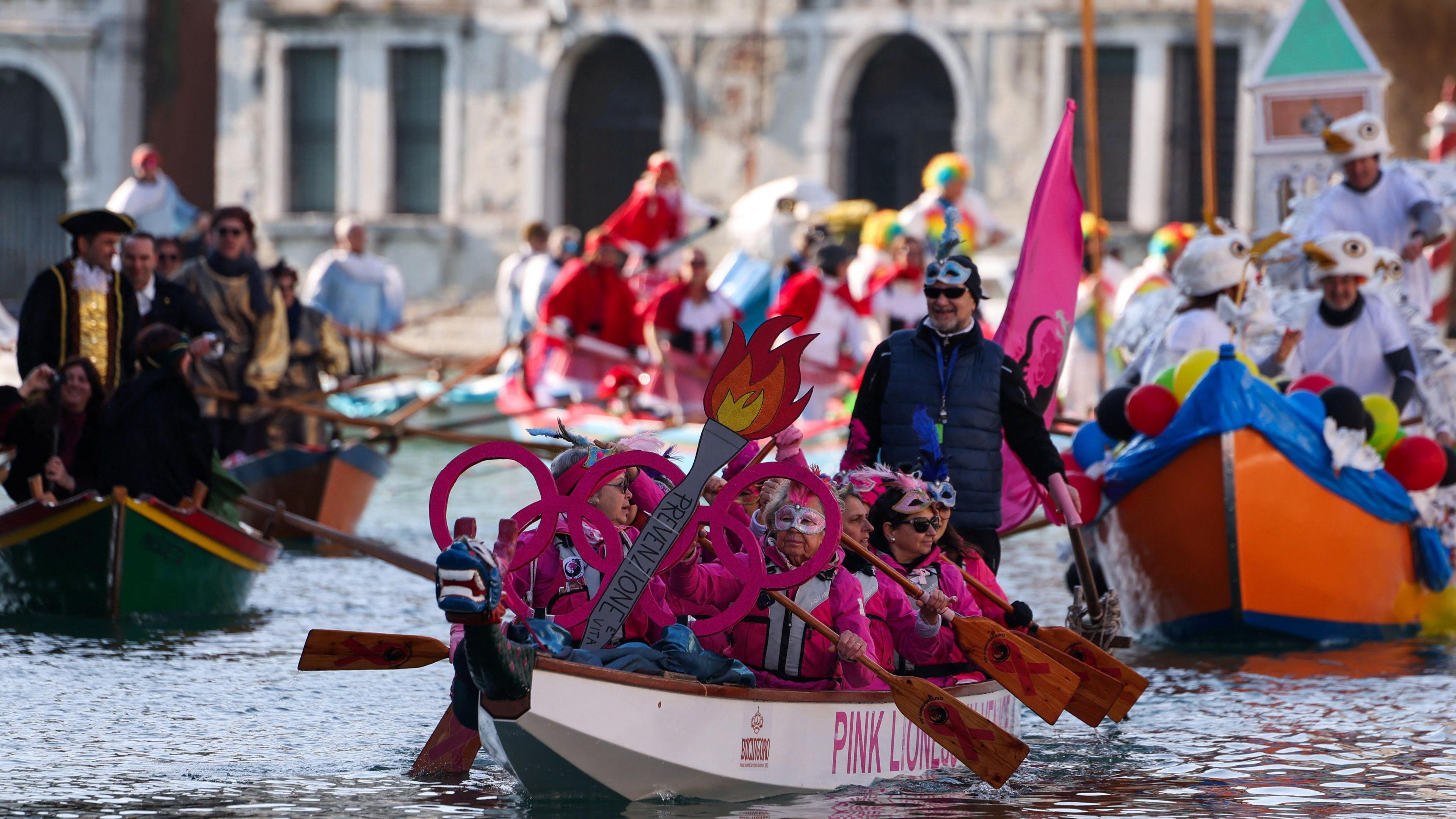 A group dressed all in pink and with glittery eye masks are pictured in a boat, with a painted cardboard torch shown on the front of their boat 