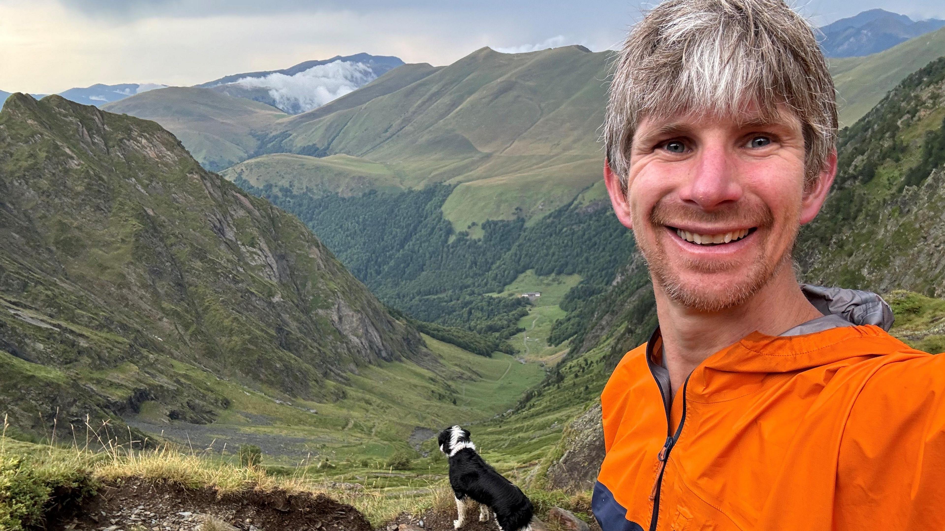 A selfie of Tim Ripper on top of a green mountain. He is wearing a bright orange jacket and his black and white collie-type dog is in the background. The hills roll off into the distance behind him with some of the furthest partially covered in cloud.