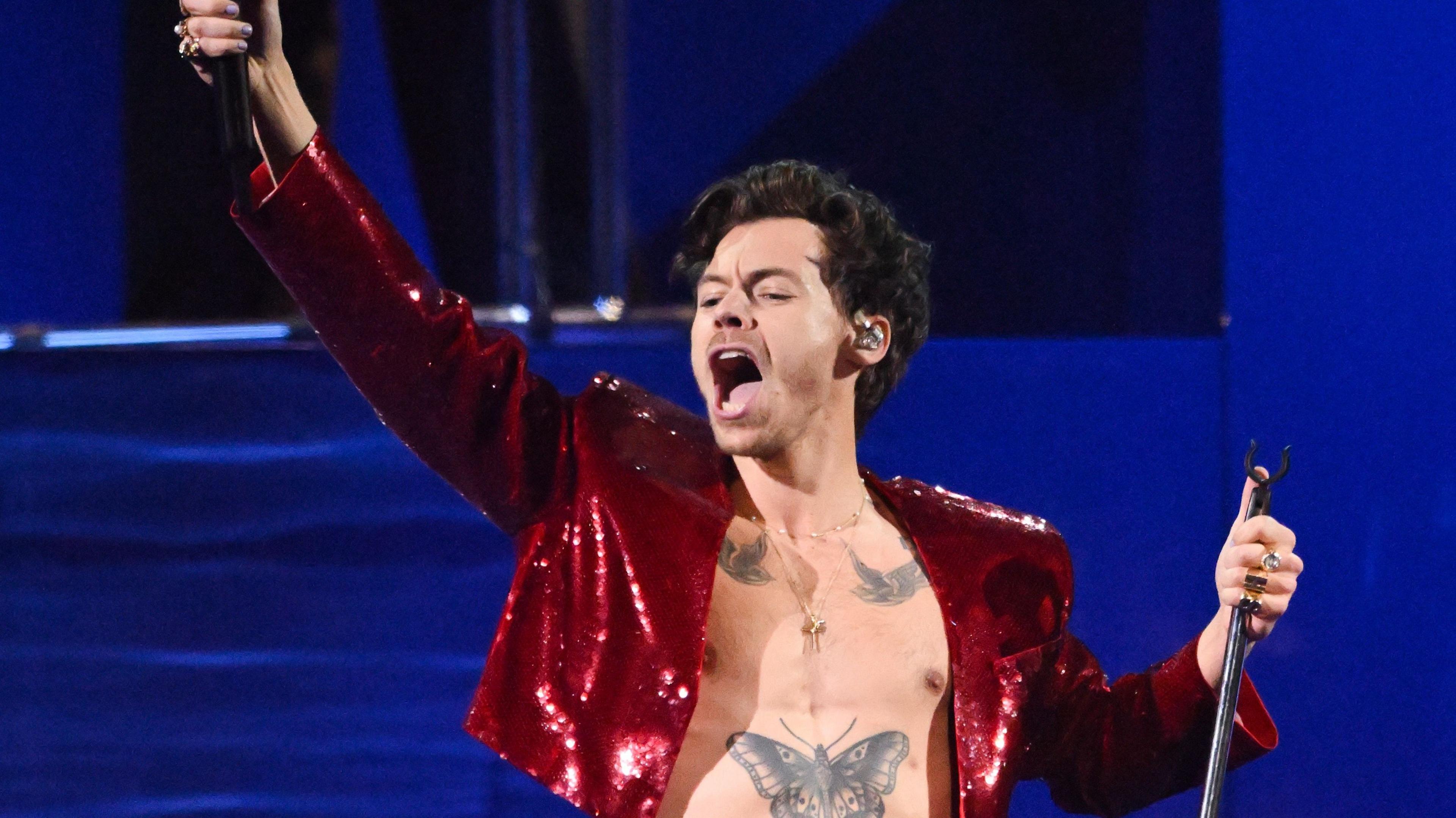 Harry Styles wears a red, cropped sequin jacket as he performs on stage in front of a dark blue background. He's holding a mic in one hand, with his arm raised in the air, and a mic stands in the other.