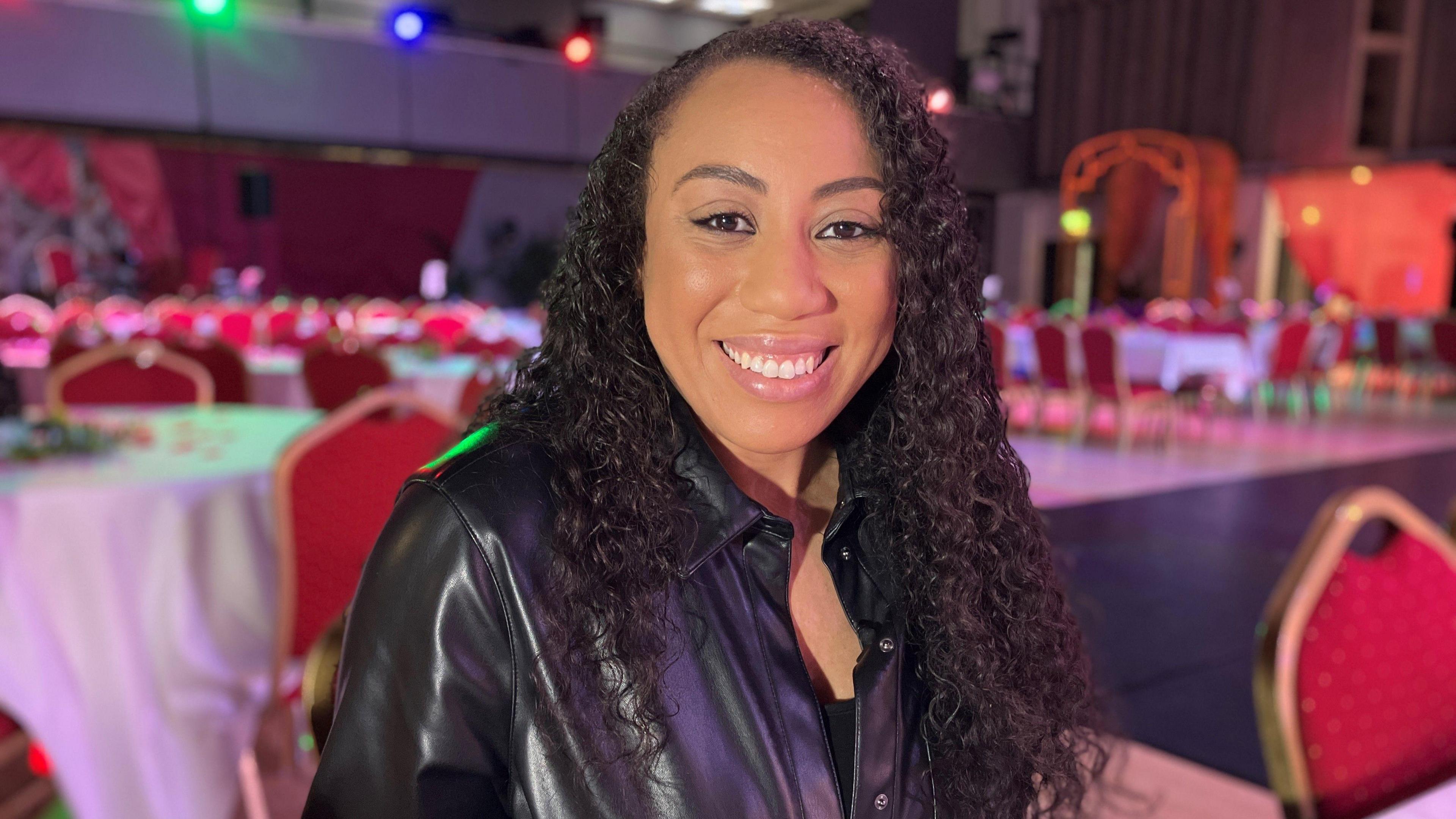 Oldham born actor Evanya Taylor with long black curly hair and a leather jacket sits at a table, with a white tablecloth, which is part of the set for The Engagement Party.