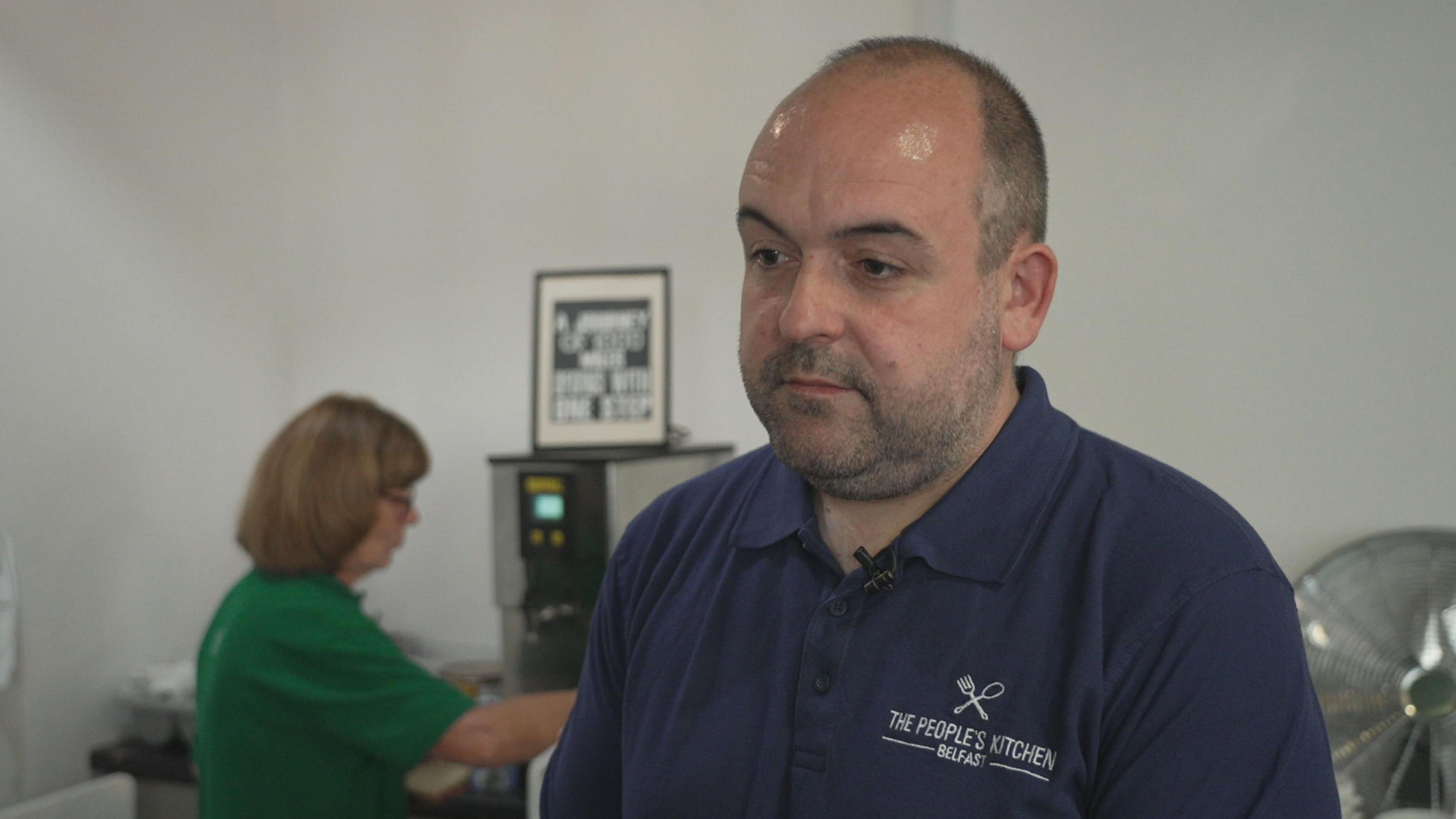 Paul McCusker, who has short, dark hair and stubble wears a navy polo shirt with the people's kitchen logo on it. Behind him, a woman in a green shirt is making tea.