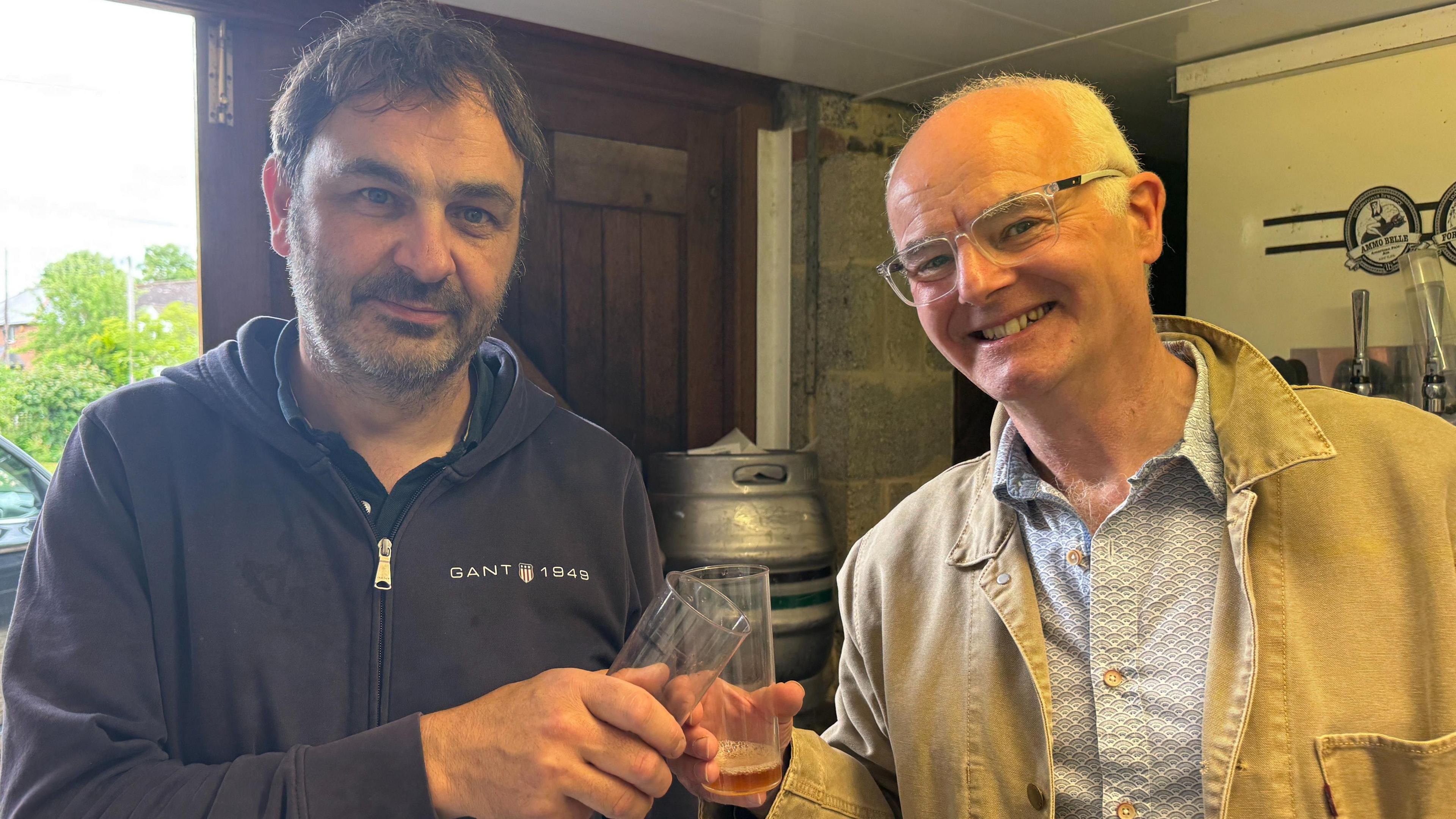 Two men pose for a photo, smiling, while clinking two glasses with a small amount of ale at the bottom. Kevin, on the left, is wearing a dark blue zipped-up hoodie and has a black and grey beard and black short straight hair. David, on the right, has white thinning hair, glasses and is wearing a white and blue patterned shirt and beige light jacket over the top. Beer kegs can be seen behind them.