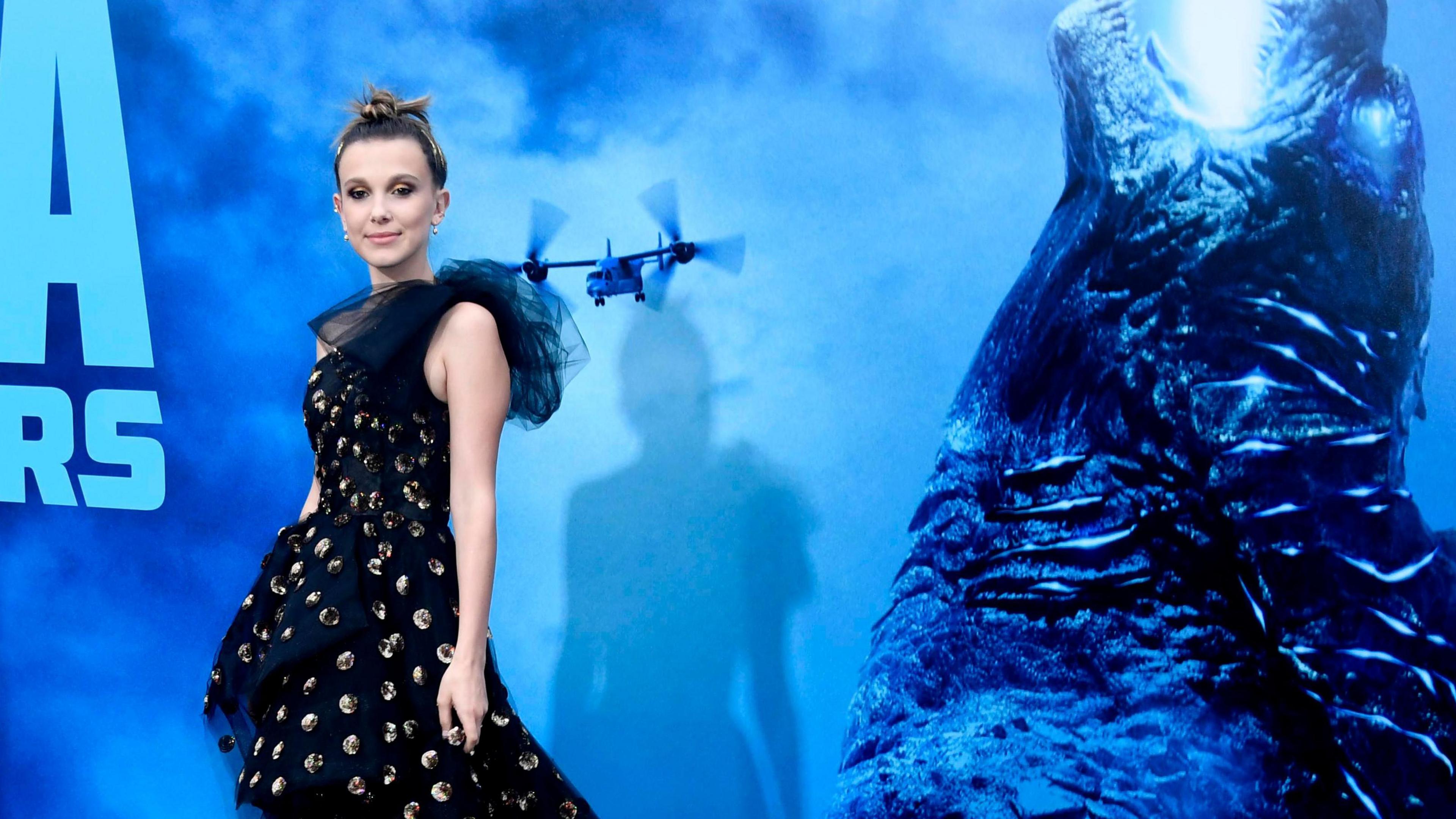 Millie Bobby Brown at the premiere for Godzilla: King Of The Monsters. She wears a long black dress with spots and ruffles on the shoulder and has her brown hair tied up in a bun