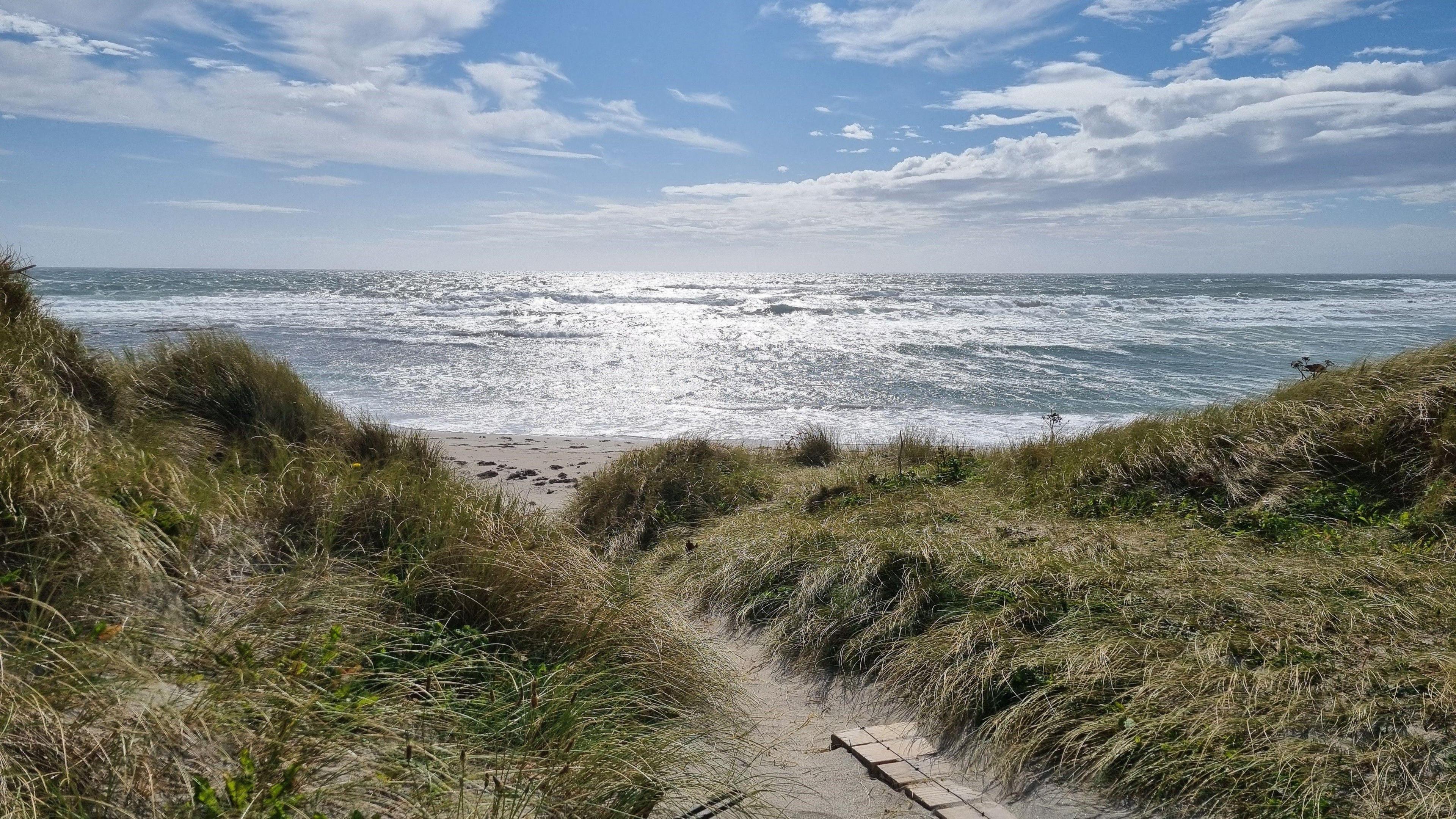 A beach in the Orkney islands. The sea is calm and the sky is blue with white clouds. A wooden walkway stretches into the sand in the foreground. There is a narrow passage between long green grass verges on either side.