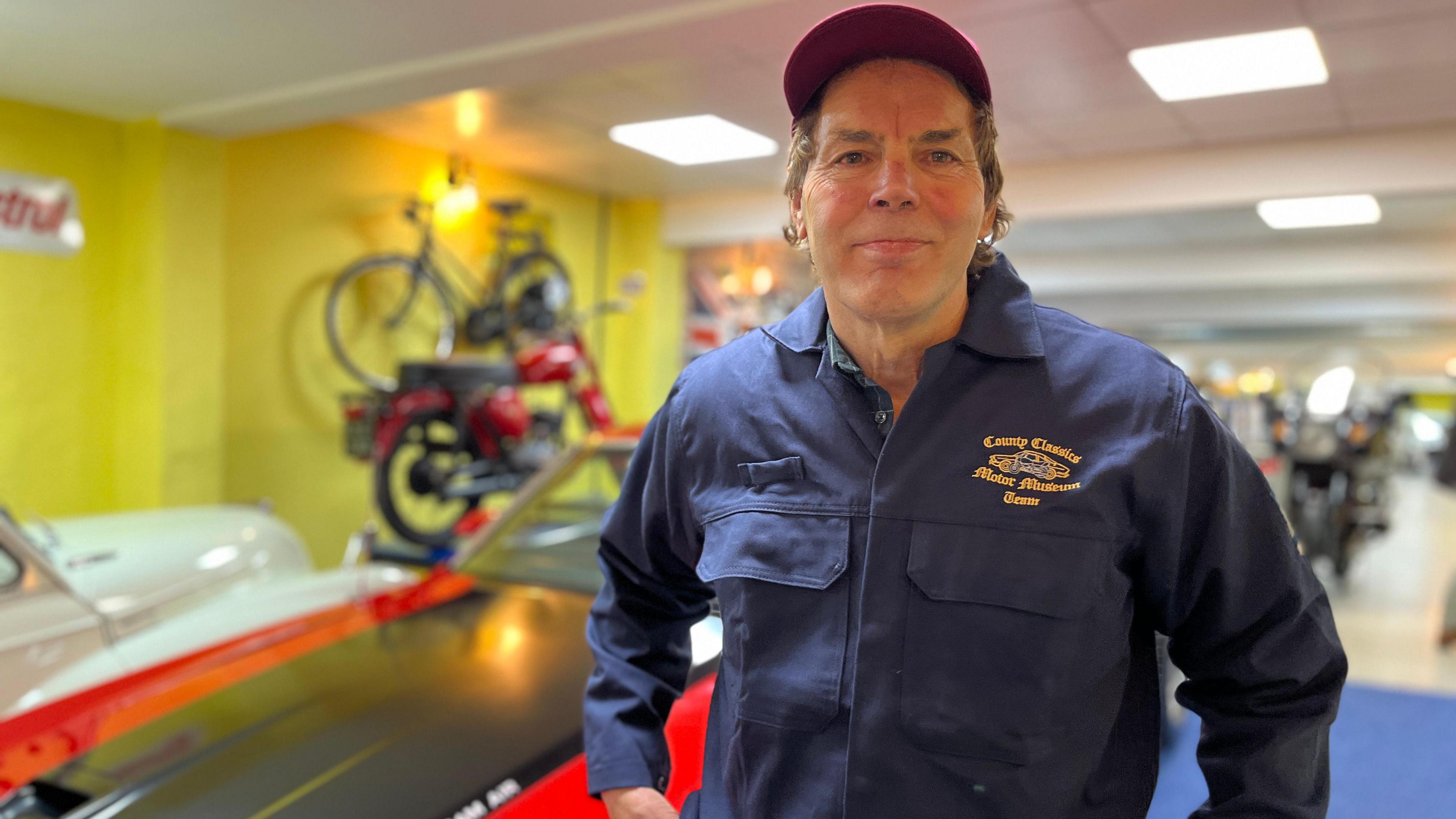Pat wearing a red trucker cap and dark blue overalls standing in his museum. A car and a motorcycle can be seen behind him but out of focus.