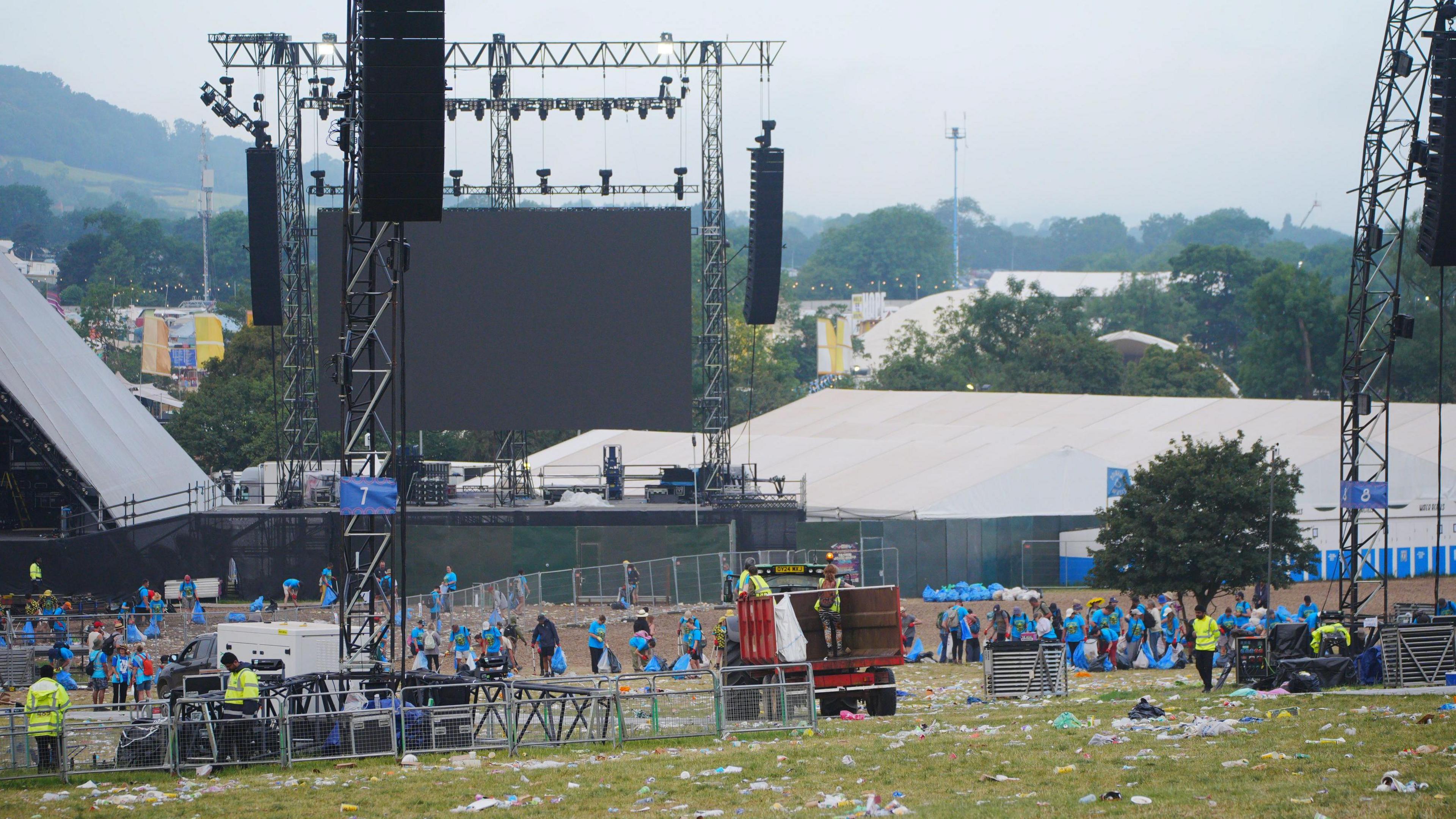 A wide shot of the Glastonbury Festival site. Part of the Pyramid Stage and the equipment used during performances can be seen. There are many volunteers litter picking dressed in blue t-shirts. Other volunteers wearing high vis yellow jackets can be seen. 