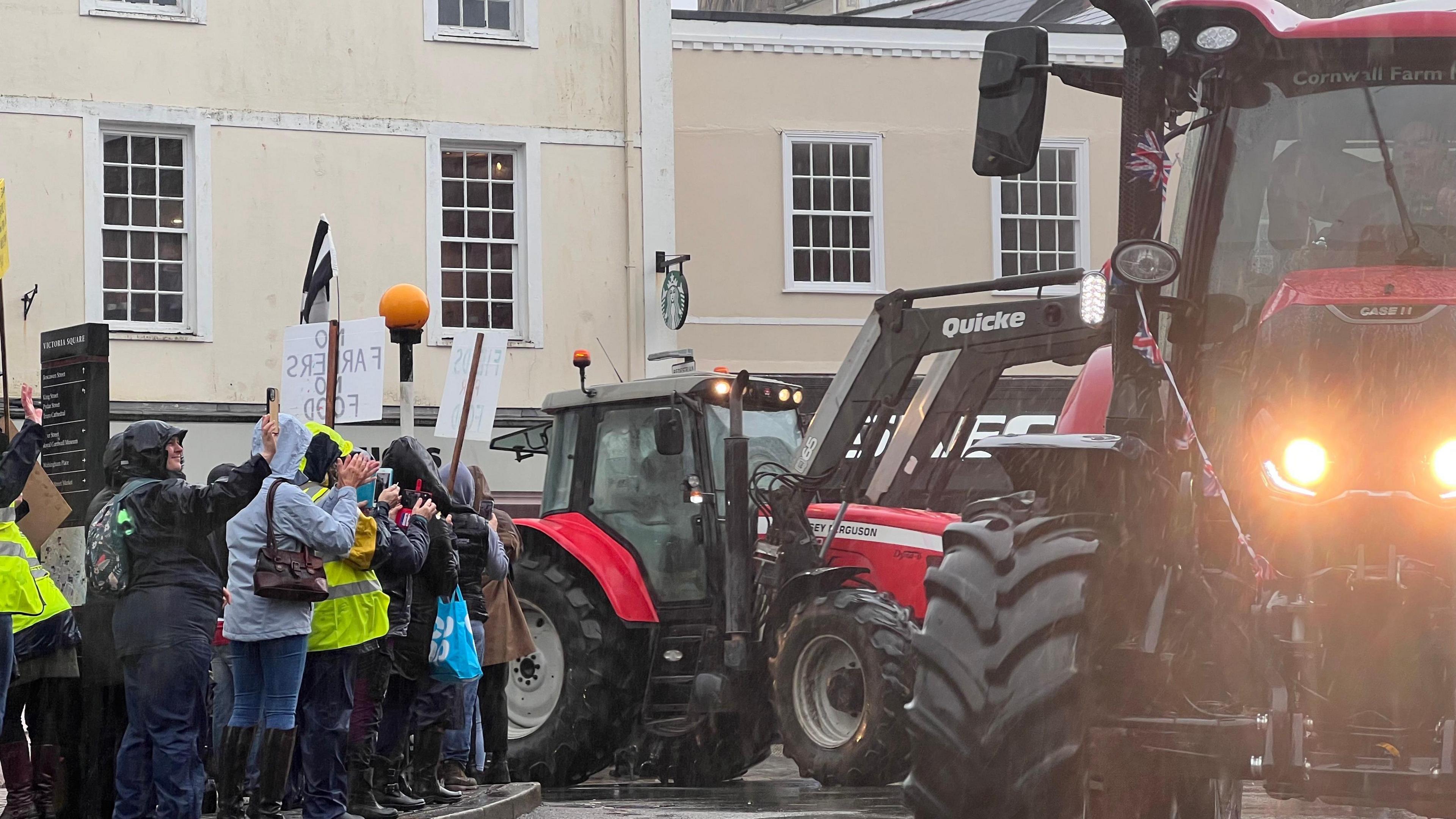 Tractors pictured in Victoria Square, Truro. They are red. There are also people stood watching the event unfold on the pavement, some holding placards