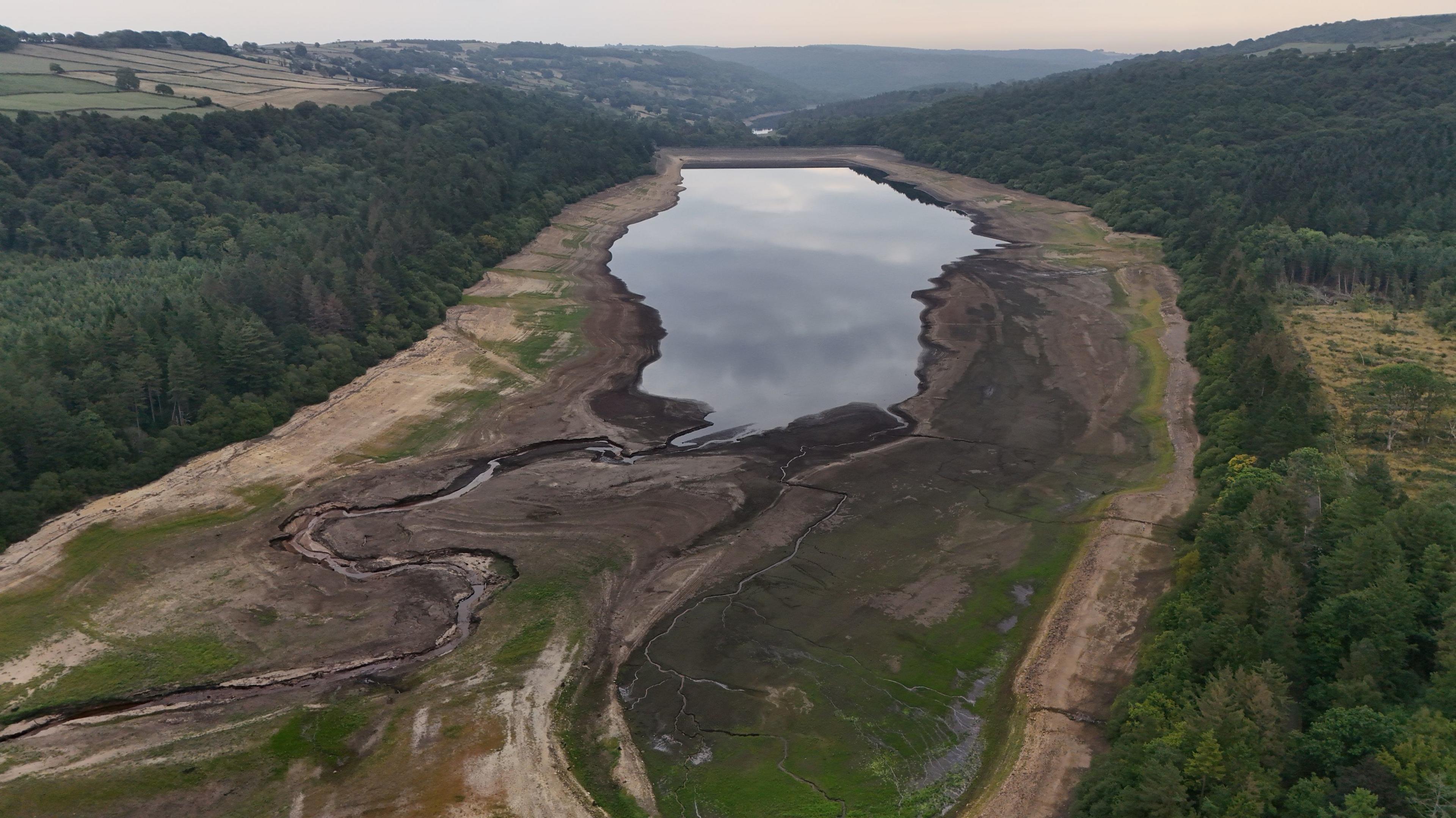 an aerial view of a reservoir, which is smaller than the area it should fill. there are grassy and sandy areas around the outskirts. On the left and right of the reservoir is a forest