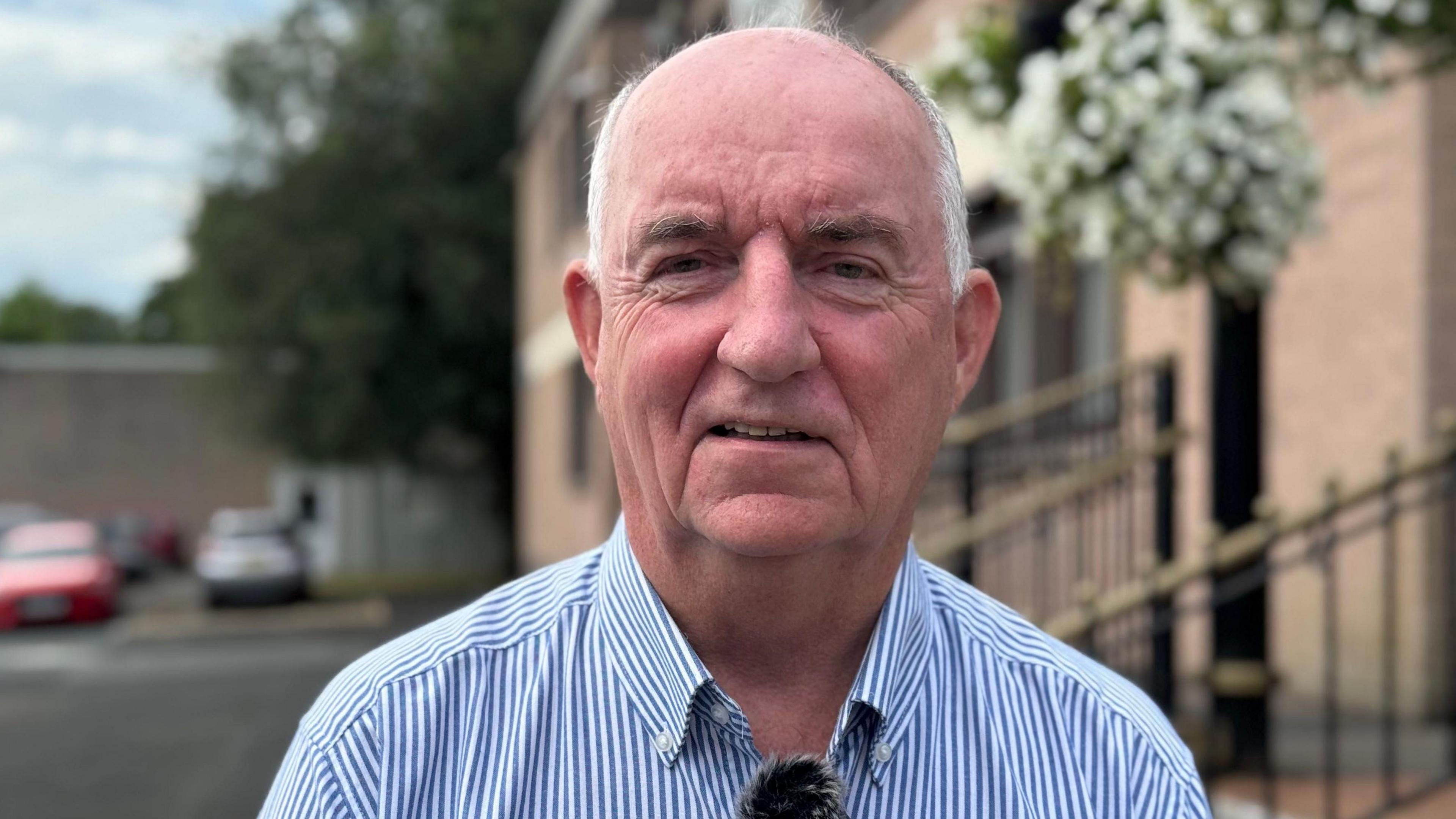 A older man looks into the camera. His hair is white around the side of his head and he is bald on top. He is wearing a blue and white striped shirt.