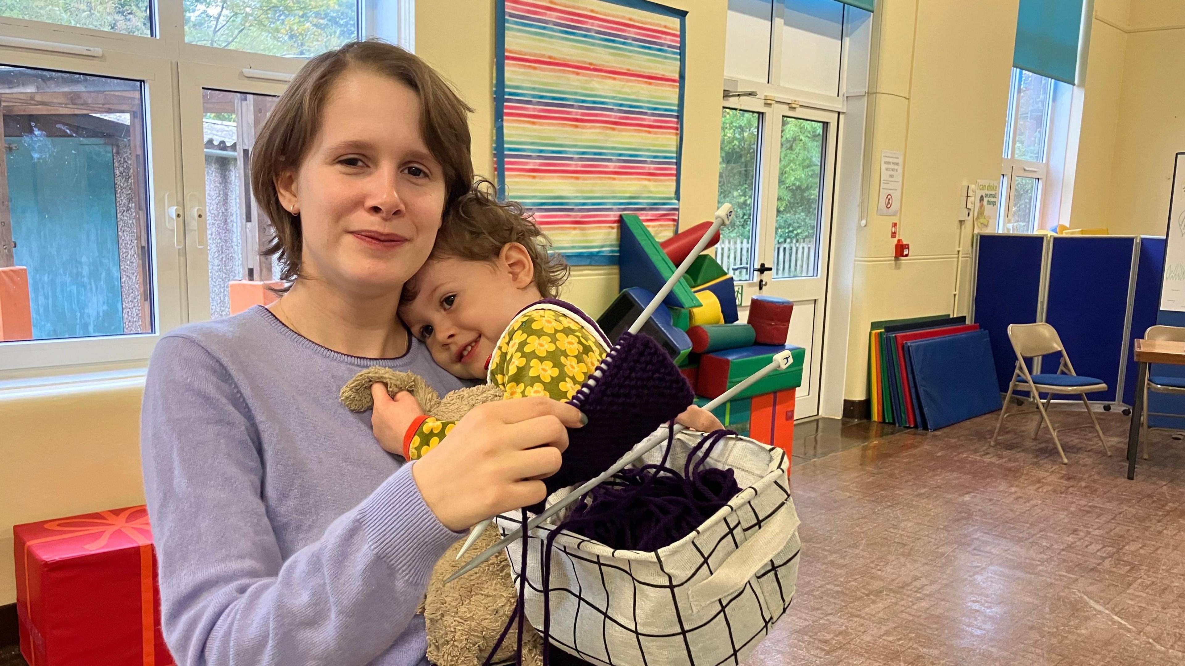 A woman wearing a purple top holding a baby and knitting materials