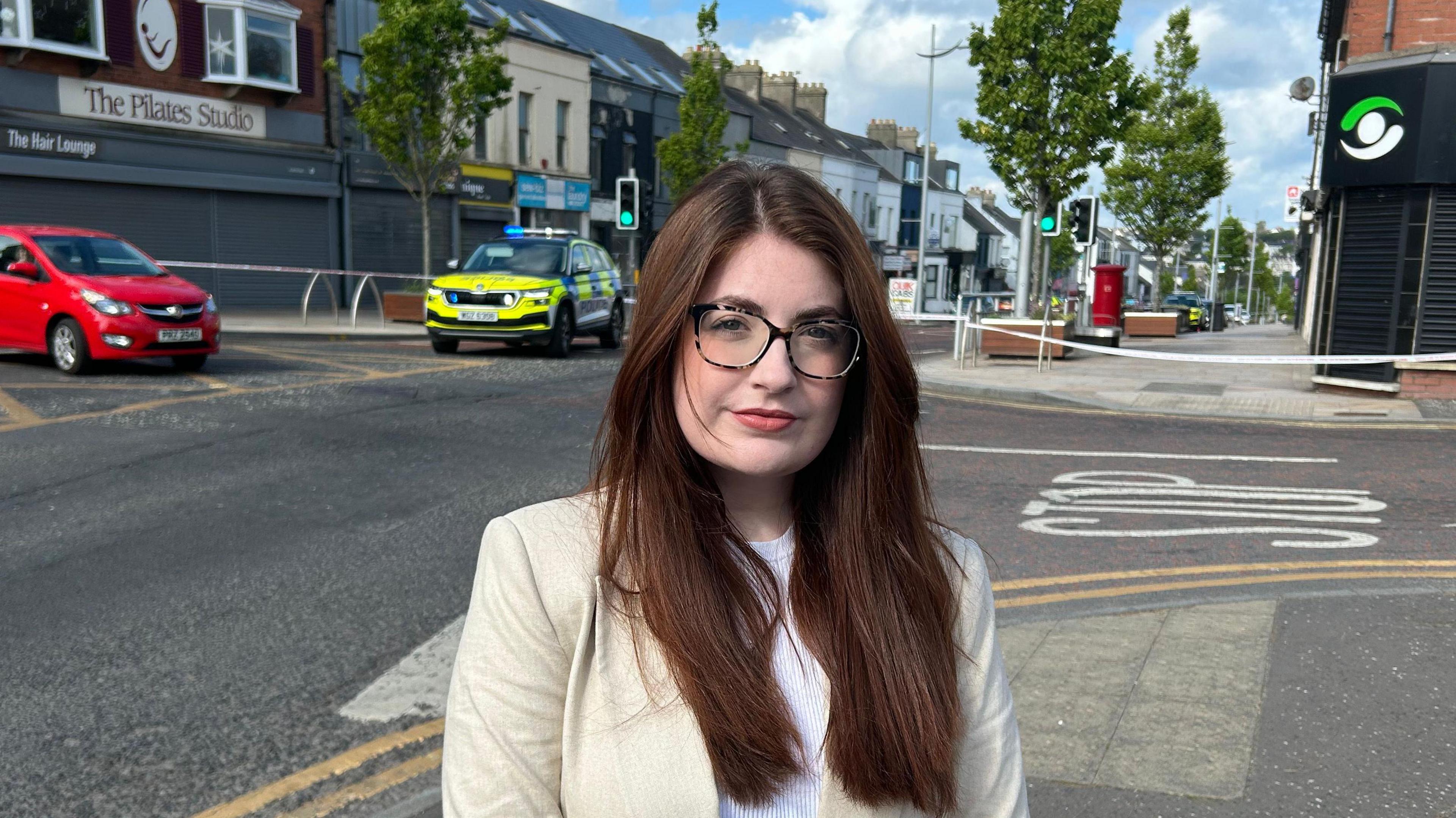 Connie is standing in front of a cordon on a high street with a police car parked beside a red car. She has long brown hair and dark glasses.