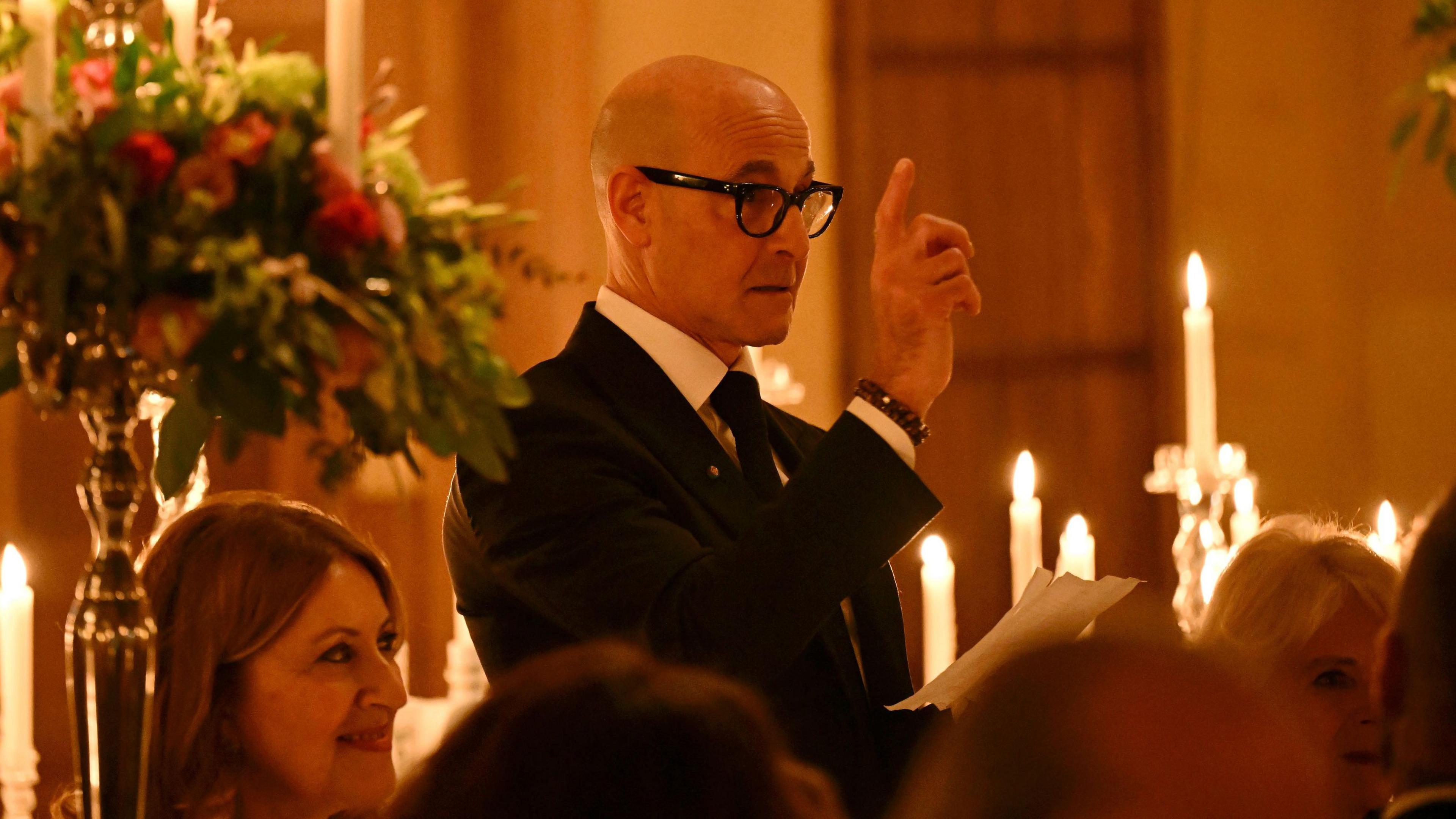 Celebrity chef Stanley Tucci, wearing a black suit and tie, gives a speech to a large group of people at Highrove House. The room is illuminated with candlelight