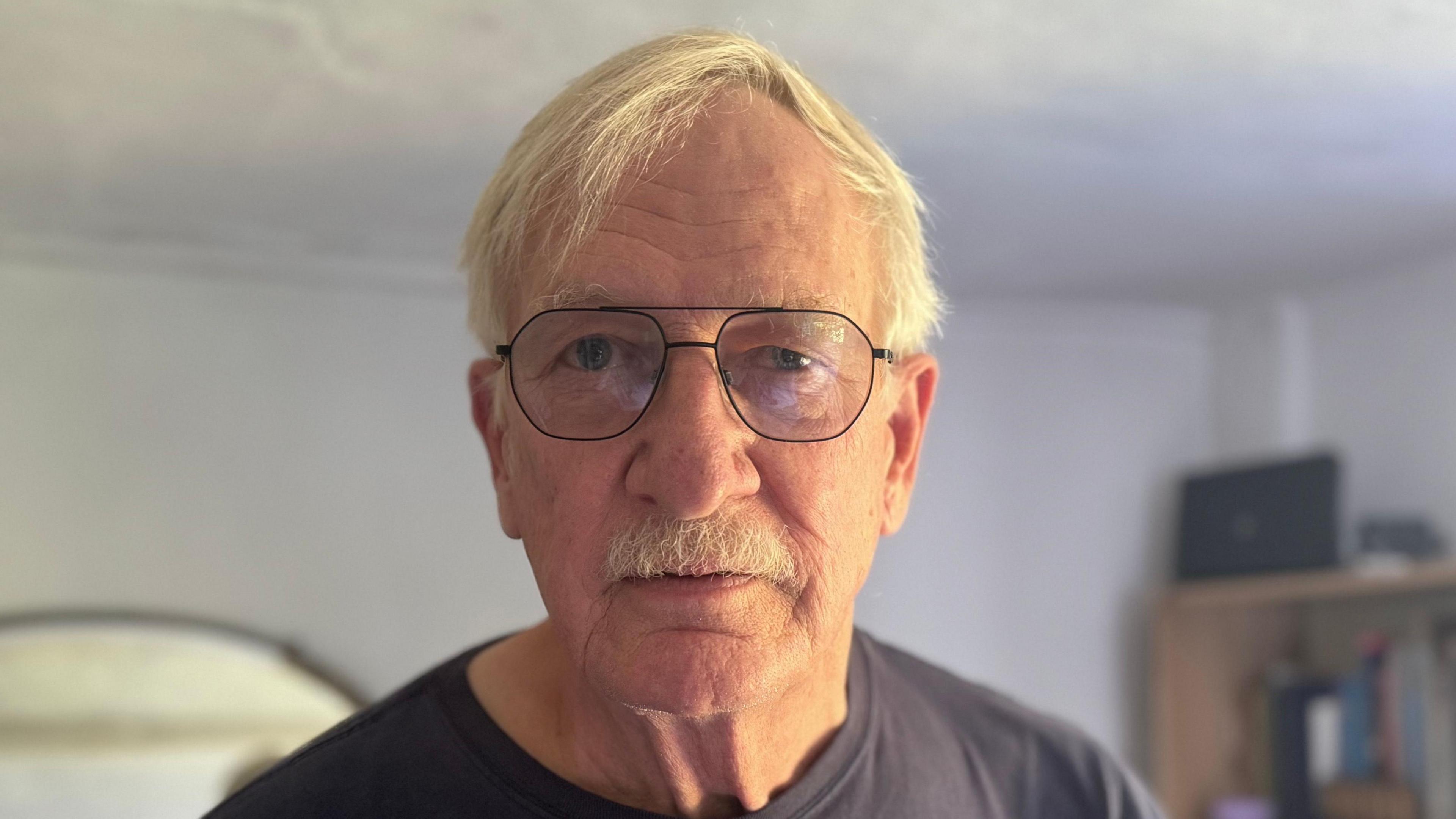 An elderly man with grey hair and glasses staring into the camera 