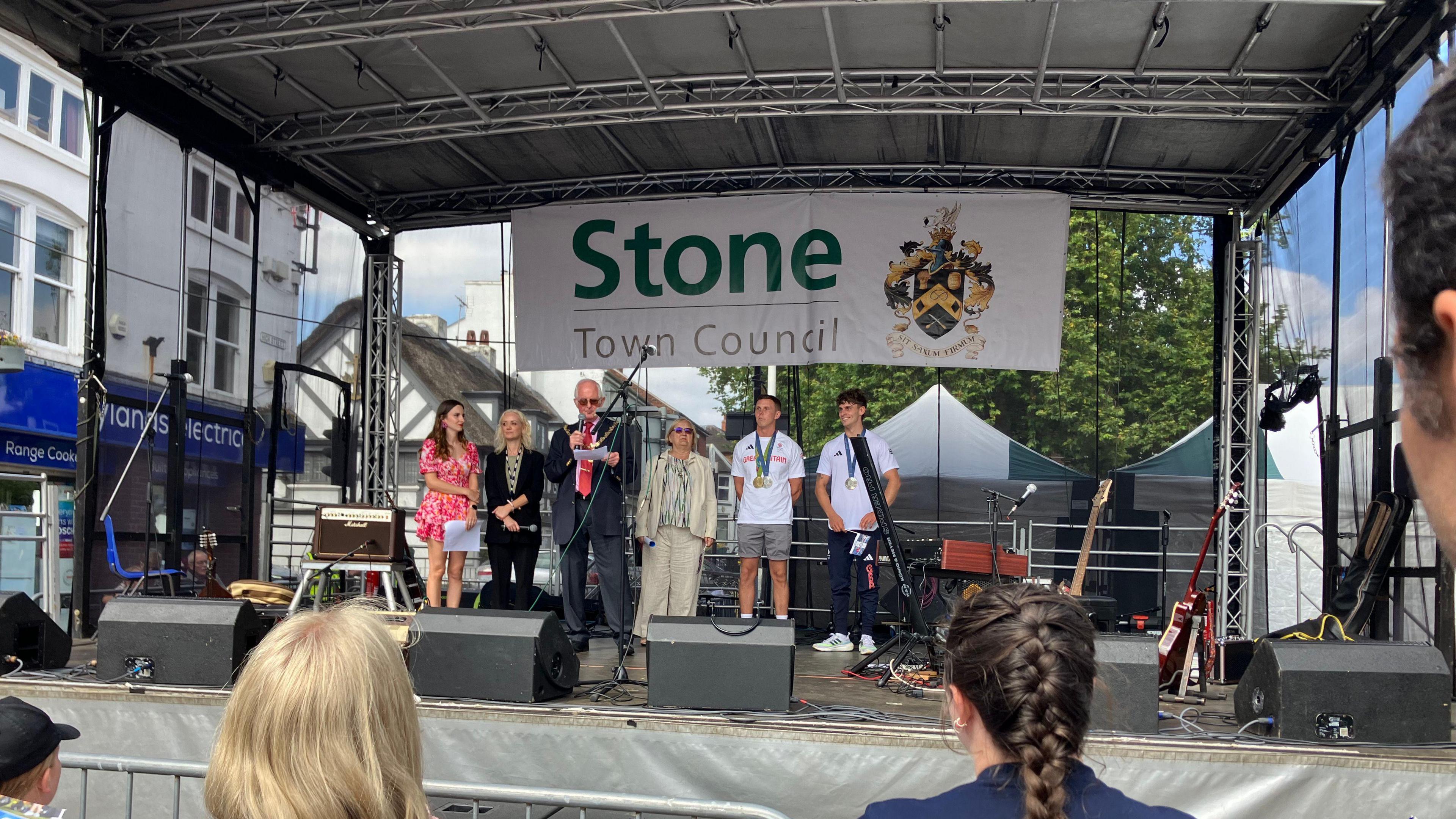 Two men stand on stage with Olympic medals around their necks. A mayor is on stage with them, speaking into a microphone. Also on the stage are three women. There is a large banner behind them that reads "Stone Town Council"