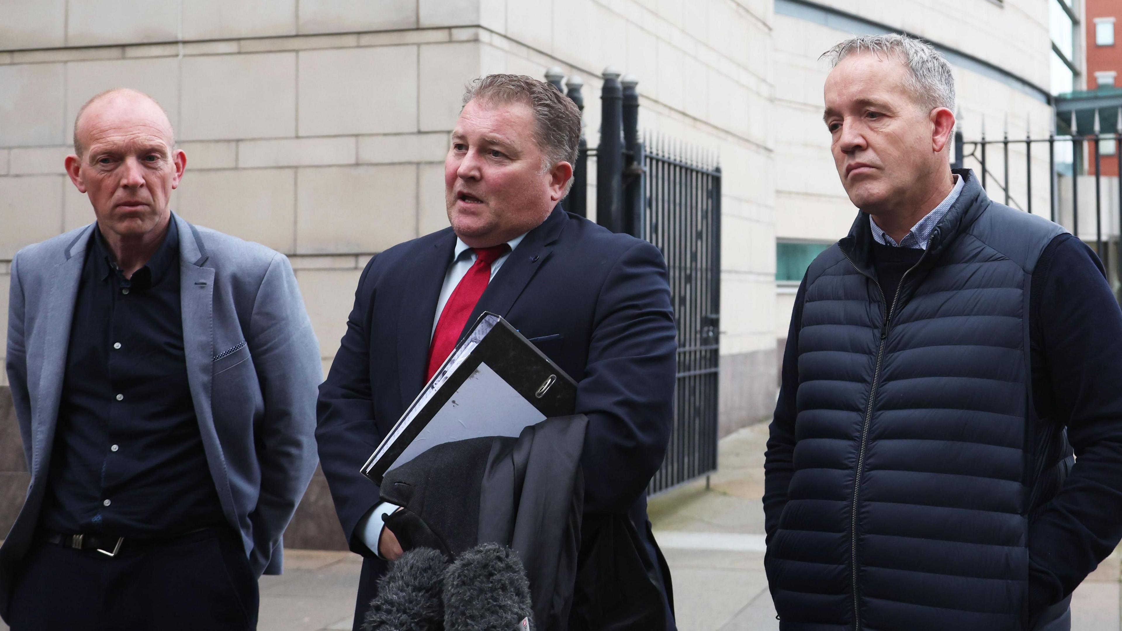 Photo outside court building showing three men in front of a microphone. Ciaran Shiels, centre, is wearing a navy suit and red tie.