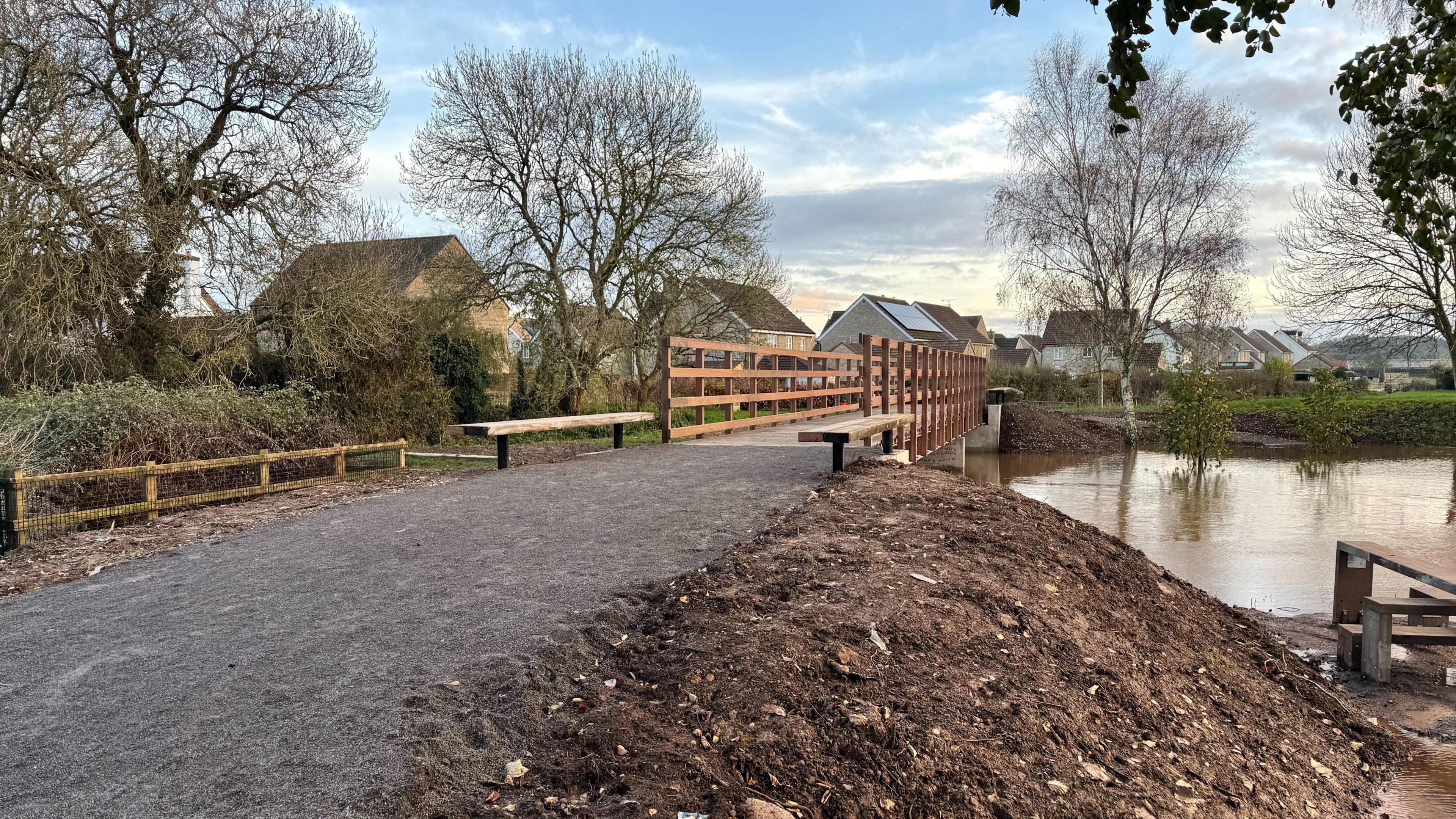 New bridge crossing a very full and overflowing river. There's lots of mud and gravel building up the pathway to a height so it can cross the river. There's a wooden bridge with houses in the background.