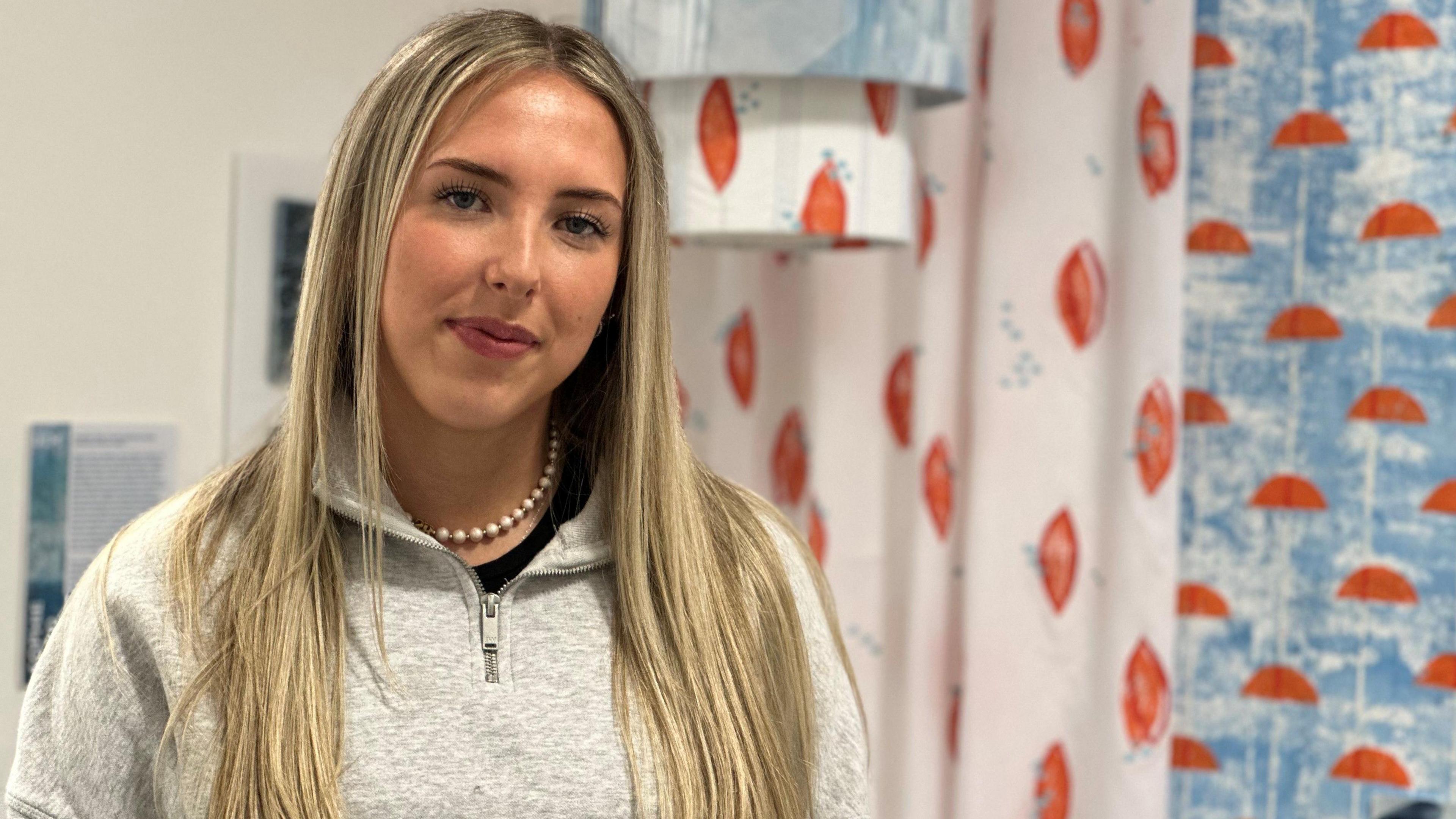 Sophie Boyde has long blonde hair and is wearing a grey top standing next to blue and orange designs on fabric.