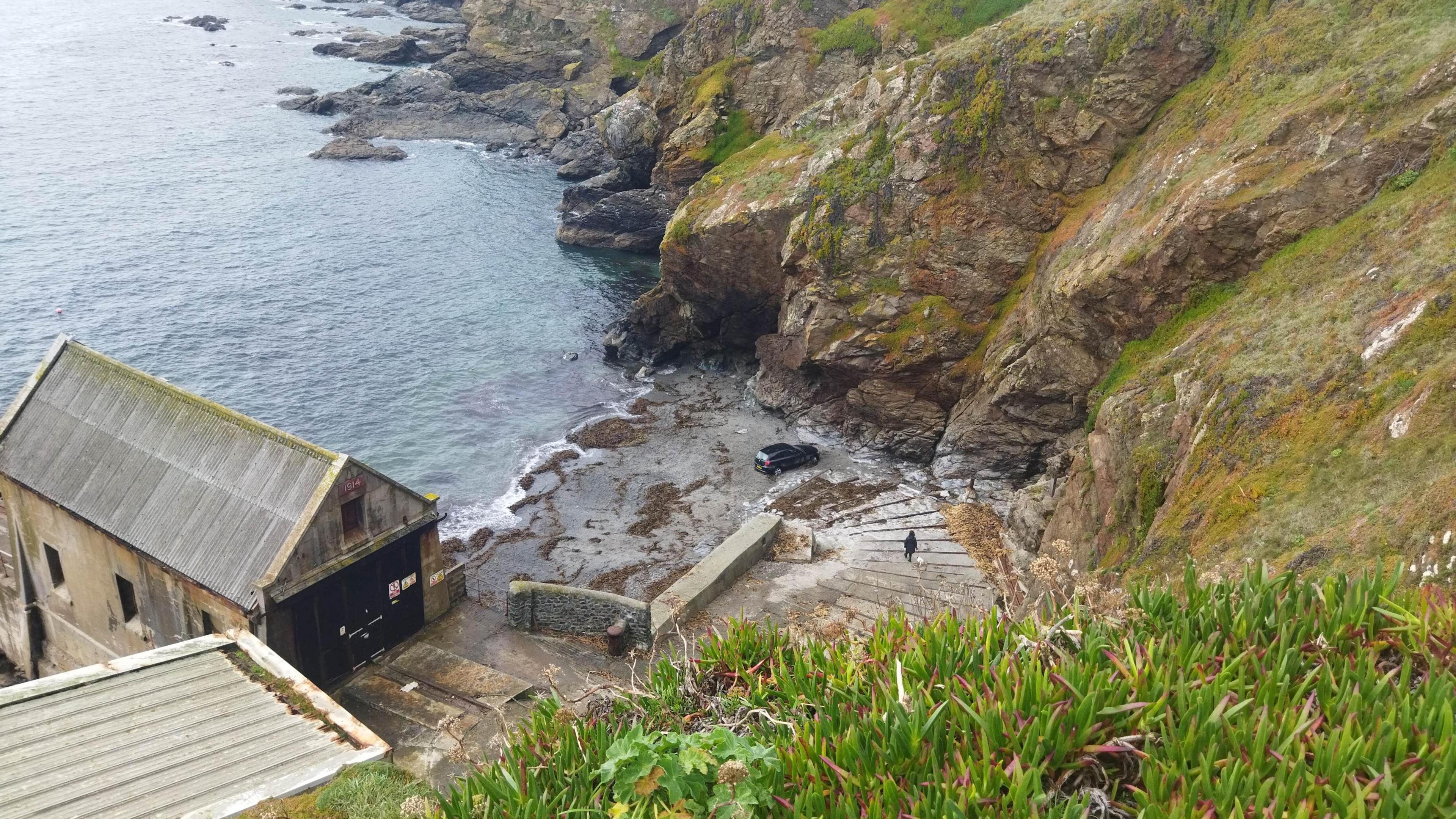 A photo of Polpeor Cove taken from the top of the cliff. A black BMW car is parked on the beach about a metre from the steep rocky side of the small cove about 10 metres from the sea. A steep stone slipway leads from the top of the cliff down to the beach below. There is an old lifeboat station on the left of the cove.  