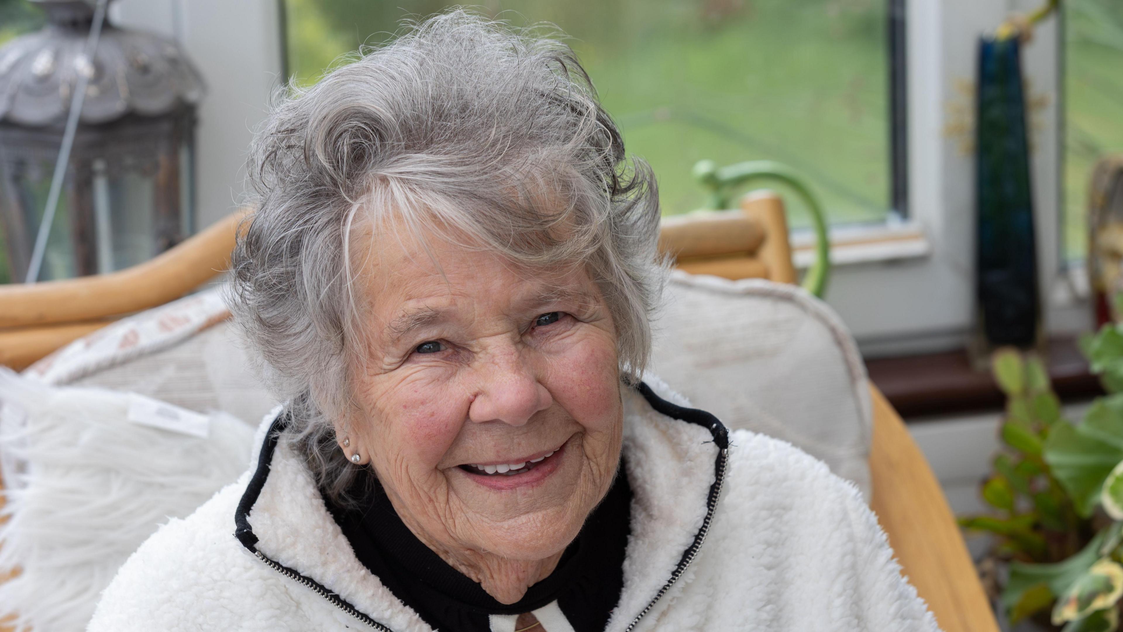 A close up shot of a smiling woman with greying hair sitting in front of a window
