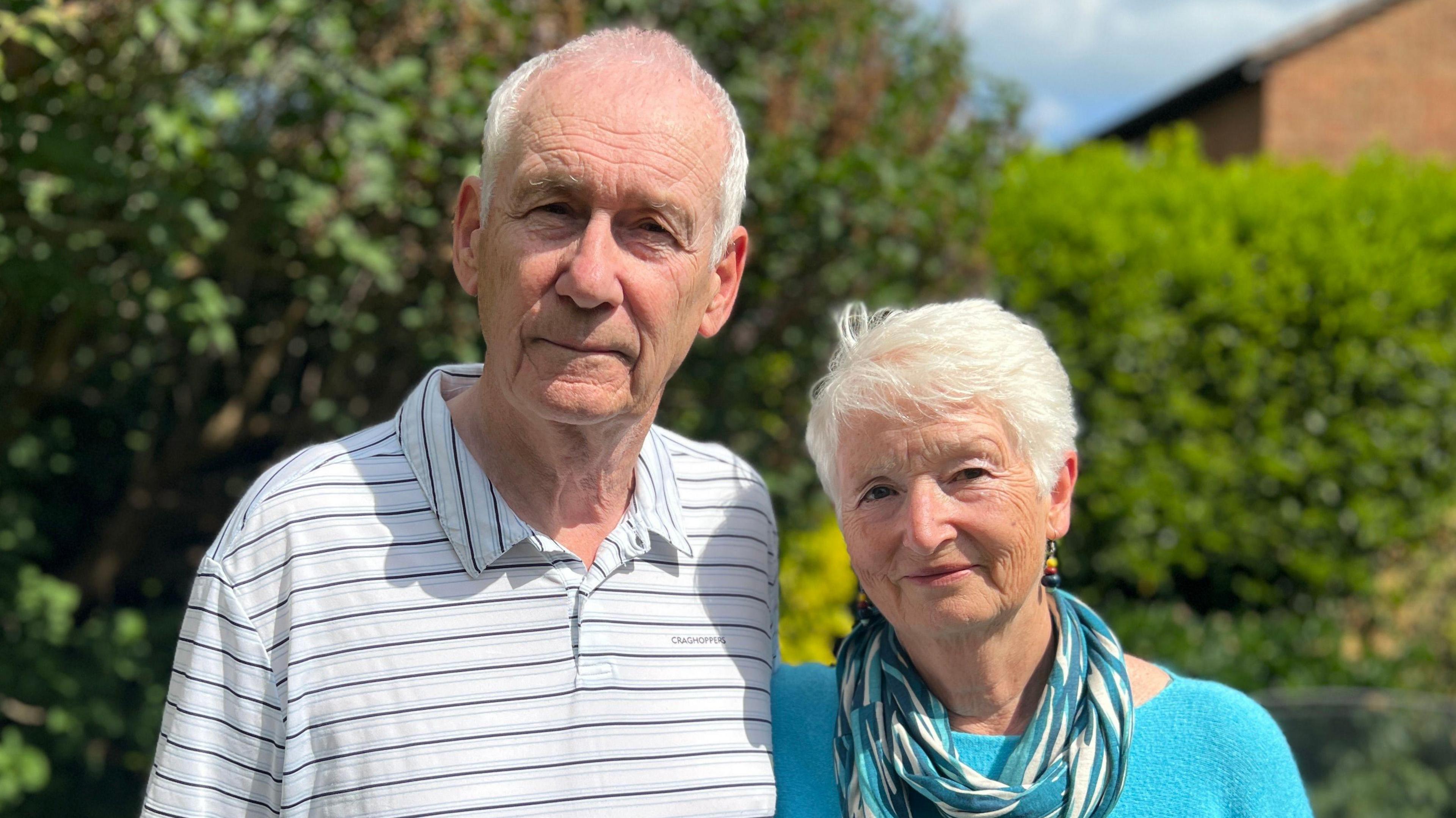 Brian Millin stands on the left with partner Maggie Reeve on the right. Brian is wearing a white polo shirt with black and grey stripes. Maggie is wearing a bright blue top with a blue scarf.